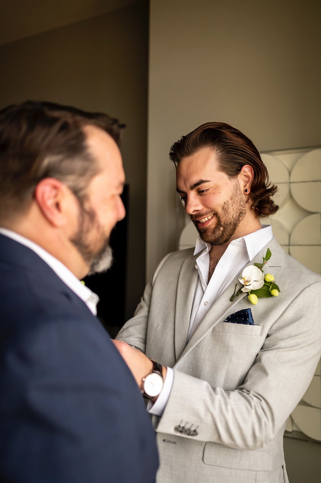 Two people in suits share a warm moment during a wedding, with one wearing light gray and a boutonniere.