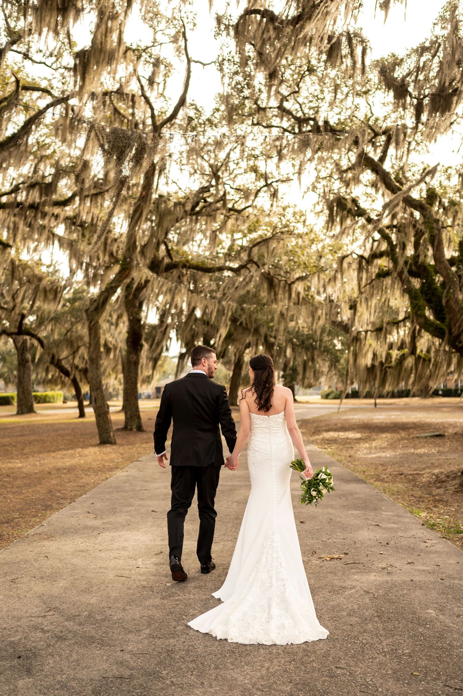 Couple walking down a path under Spanish moss draped oak trees at golden hour, bride holding wedding bouquet.