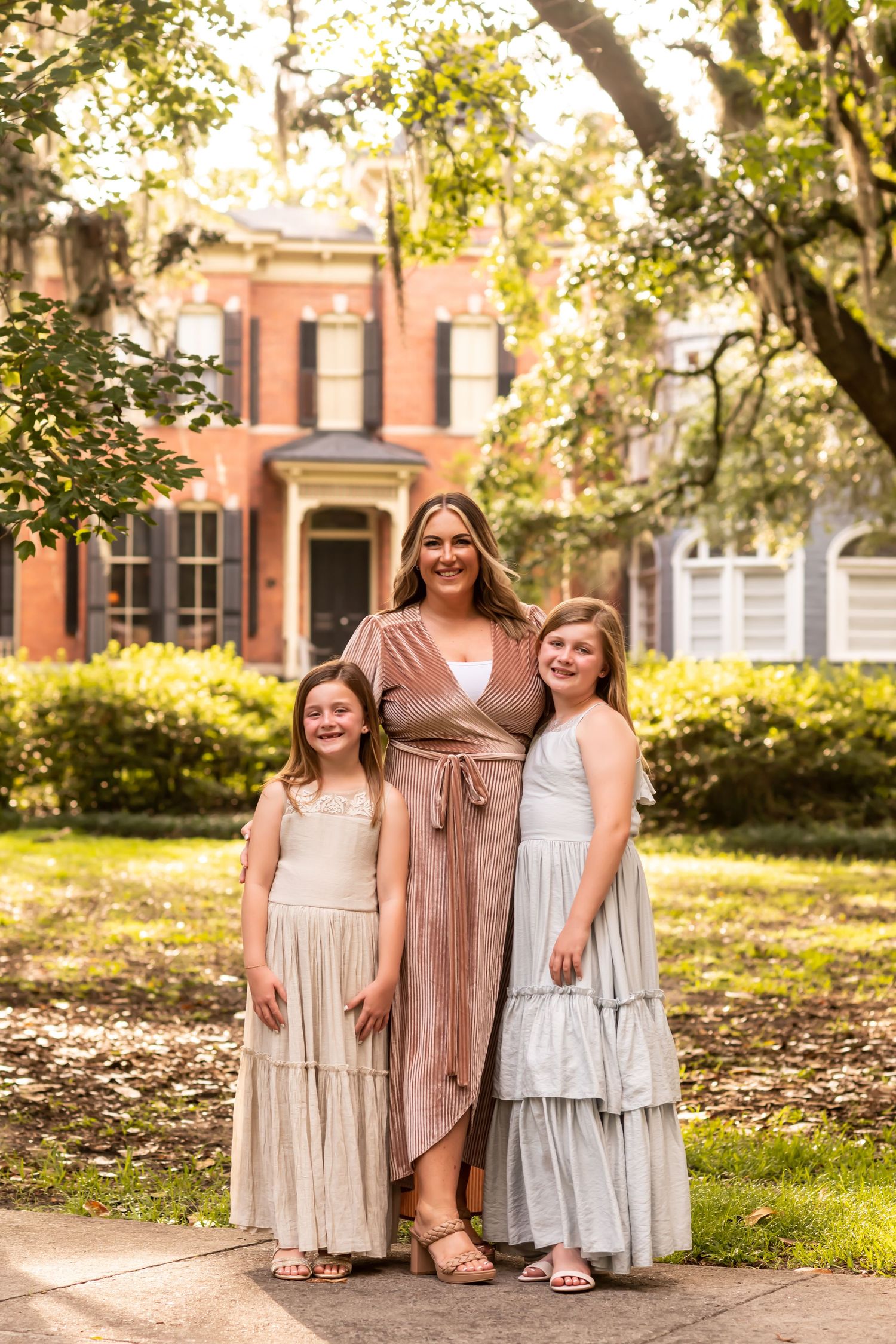 Family wearing coordinated neutral dresses poses together on a historic tree-lined street in Savannah.