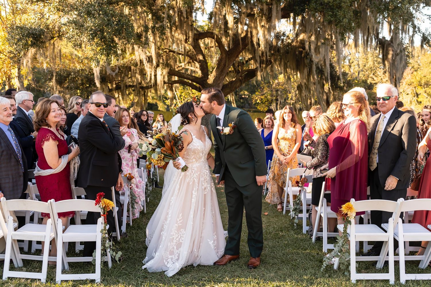 Wedding ceremony guests gathered under Spanish moss-draped trees with white folding chairs at sunset.