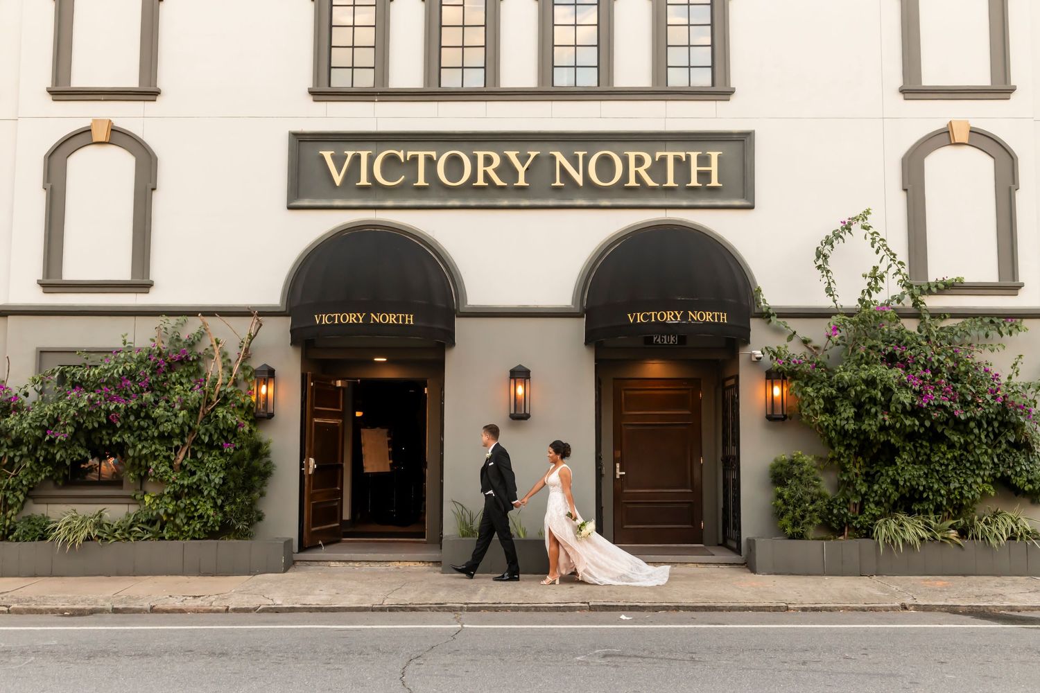 A couple stands between wooden doors at the entrance of Victory North venue with classic architectural details and greenery.
