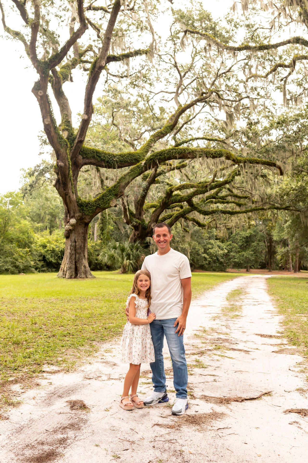 Two people stand together on a dirt path under a massive live oak tree draped with Spanish moss in a serene park setting.