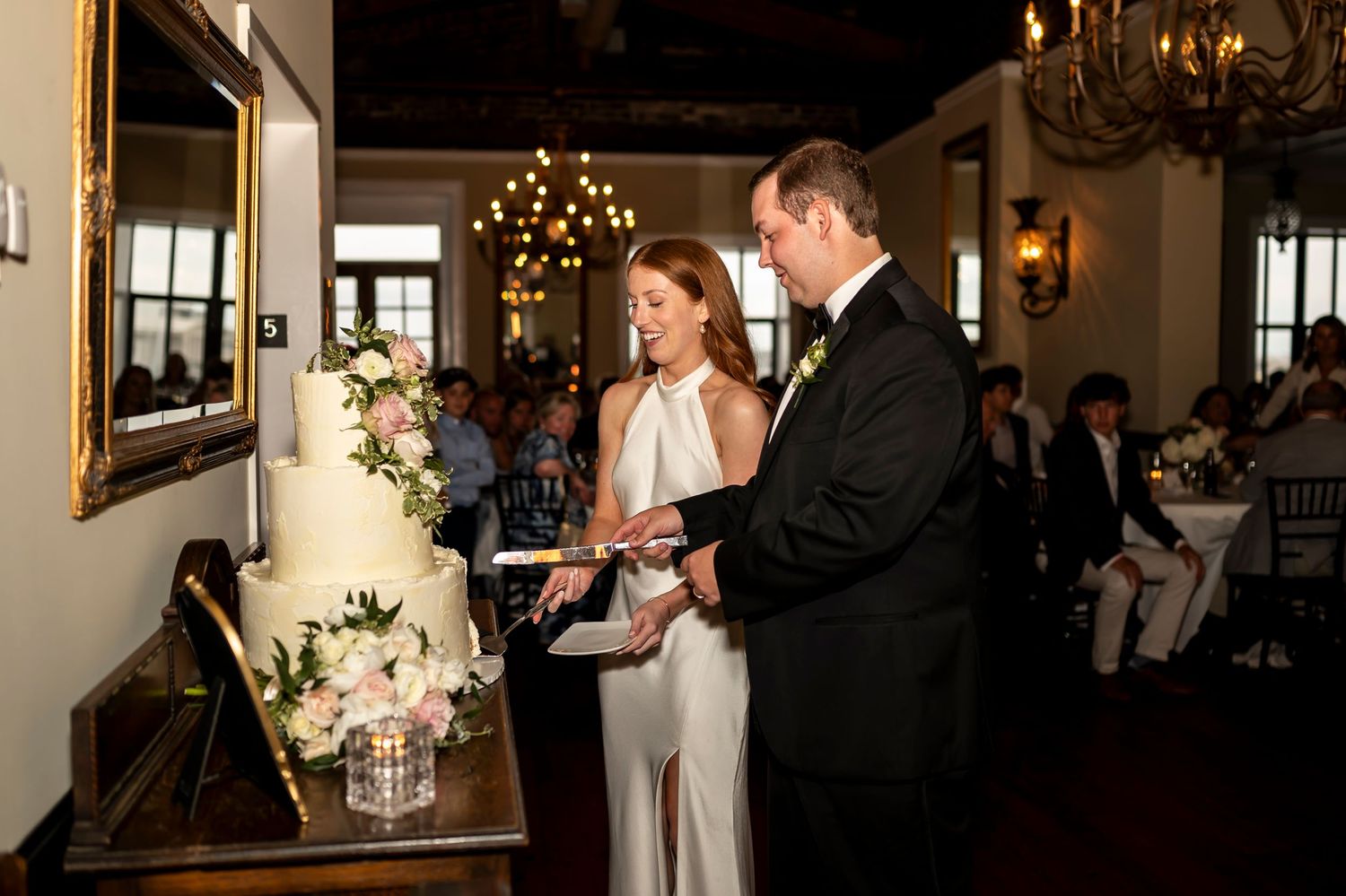 Couple cutting their elegant white wedding cake adorned with flowers in a chandelier-lit reception venue.