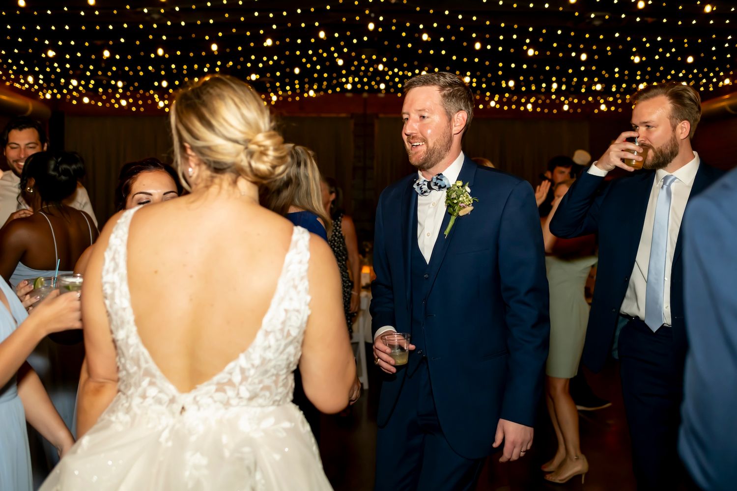 A couple dances at their wedding reception under twinkling string lights while guests celebrate in the background.