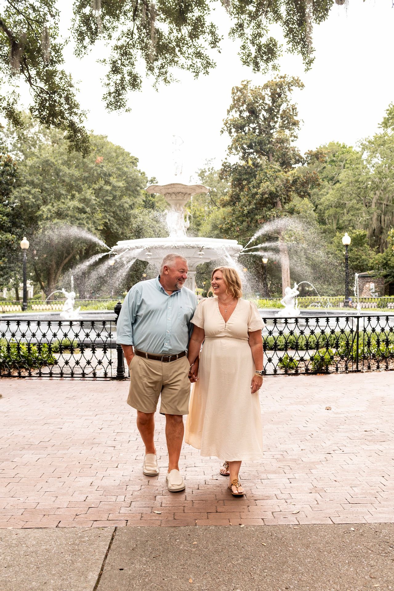 A couple takes a romantic stroll in front of a cascading fountain surrounded by lush trees.