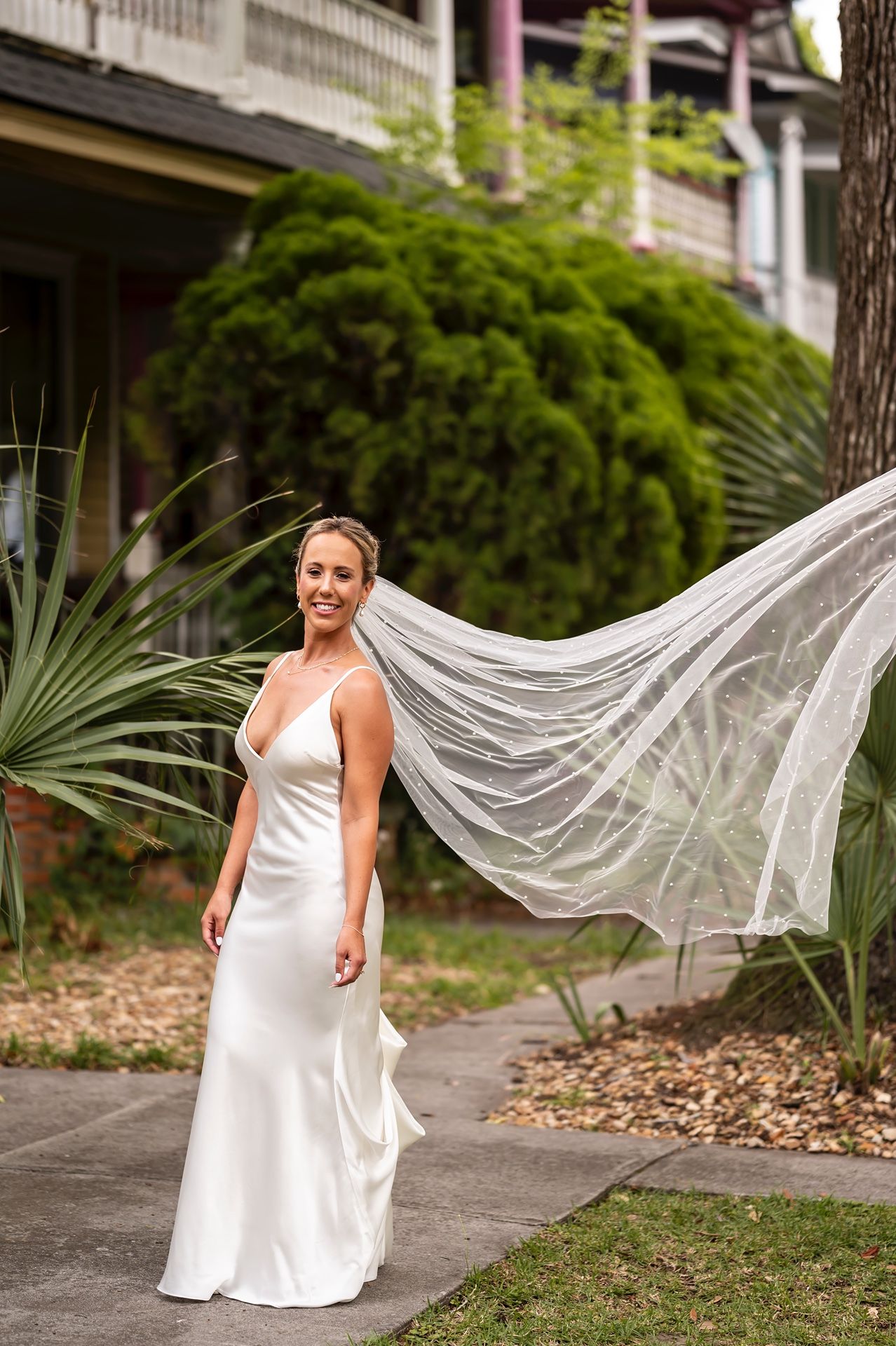 A bride in a classic white silk dress stands outdoors while her long flowing veil catches dramatically in the breeze.