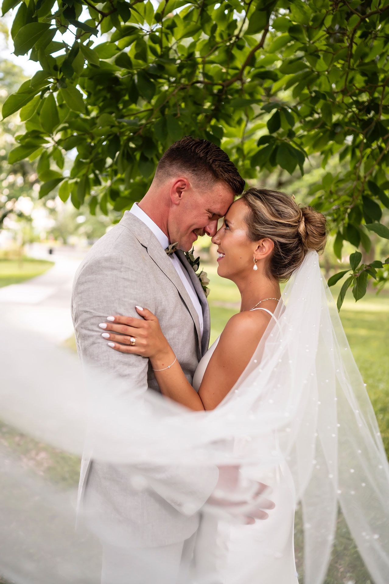 Newlywed couple shares intimate moment under green leaves with flowing white veil in outdoor garden setting.