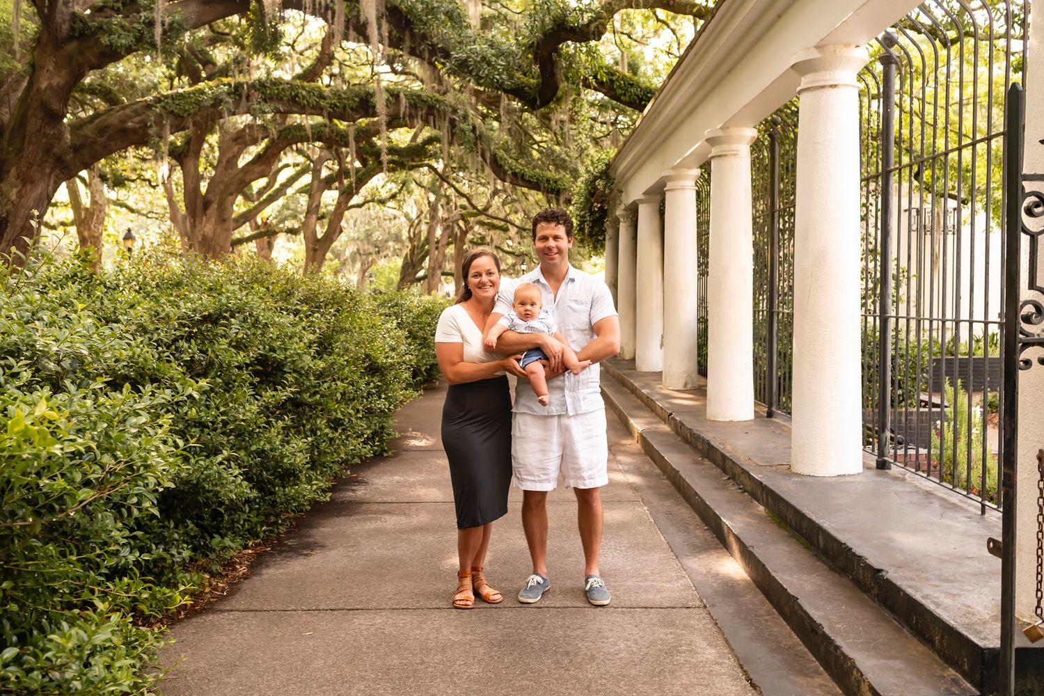 Two people walk together down a covered sidewalk with picturesque oak trees and white columns along a garden path.