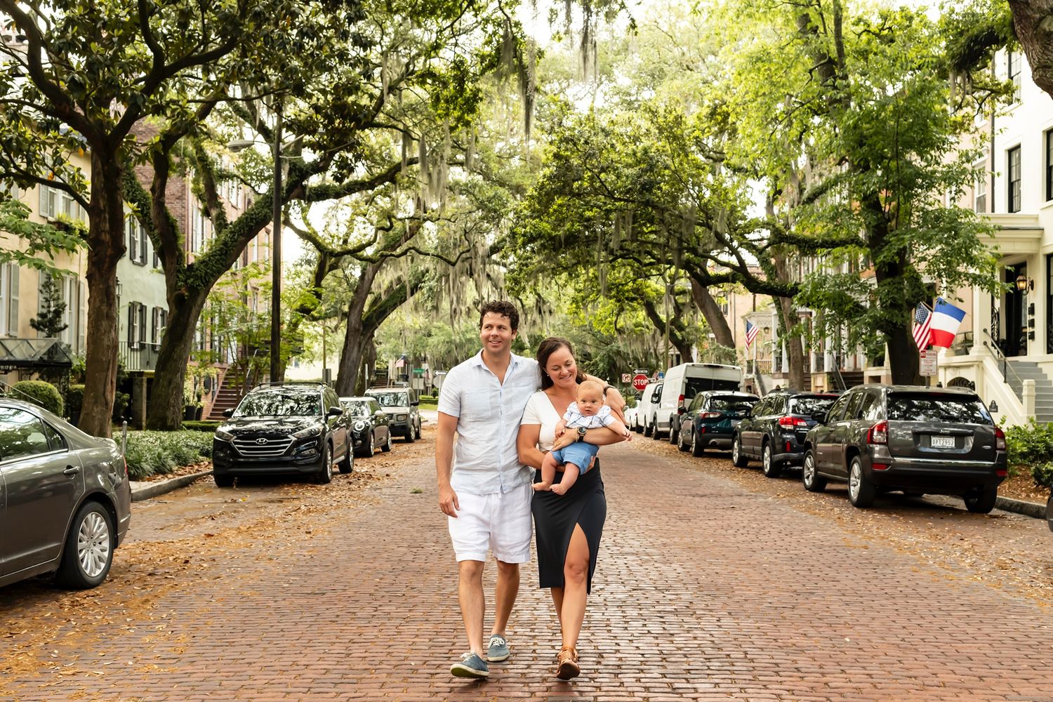 Two people stroll down a charming brick-paved street lined with moss-draped oak trees and parked cars in a historic district.