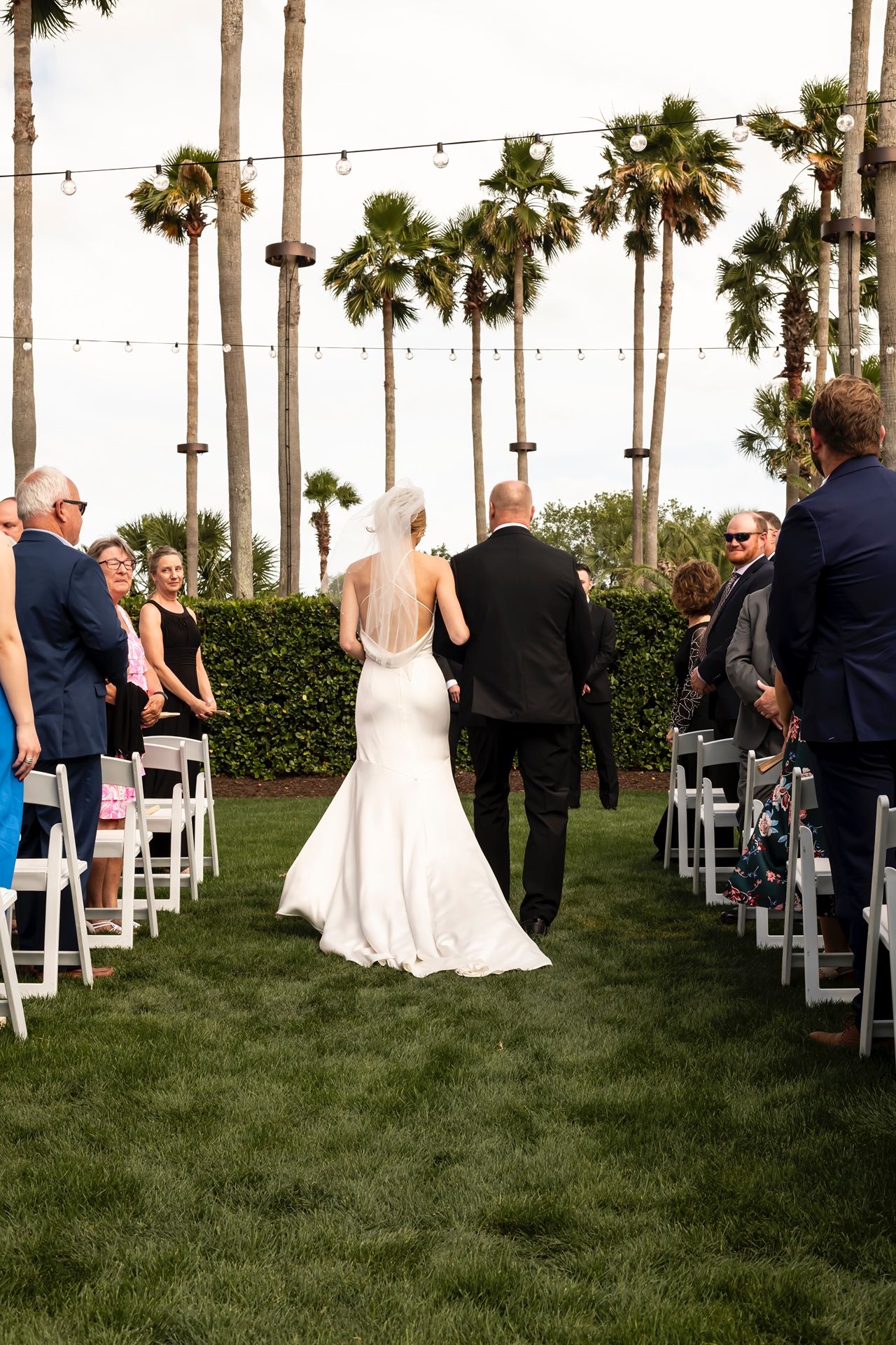An outdoor wedding ceremony under tall palm trees with string lights and guests seated on white chairs on green lawn.