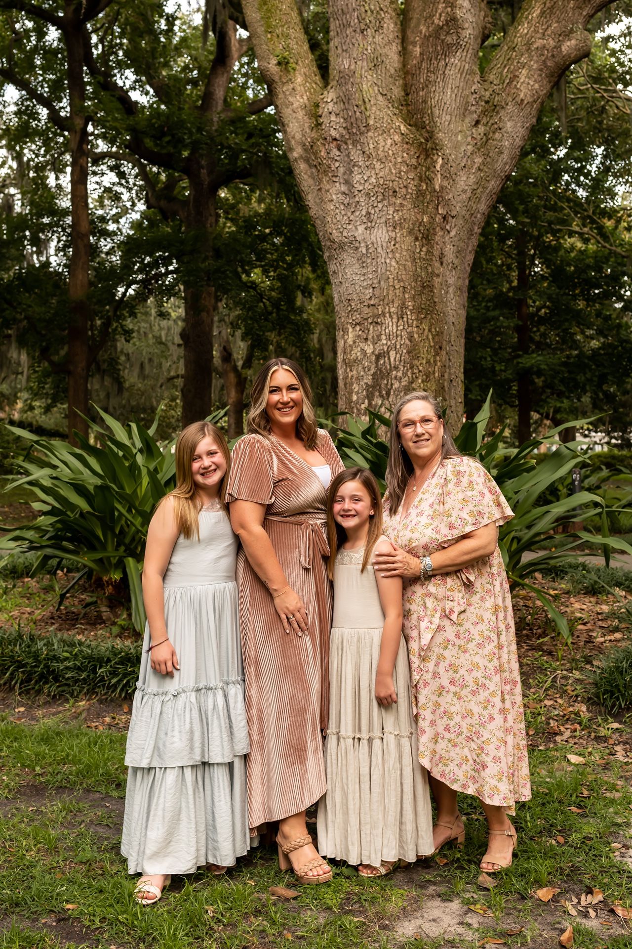 Four people in coordinating dresses pose together in front of a large tree with lush greenery in a garden setting.