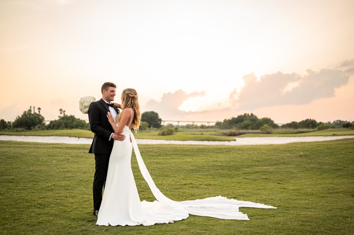 A couple embraces on a green golf course during sunset, silhouetted against the warm glowing sky.