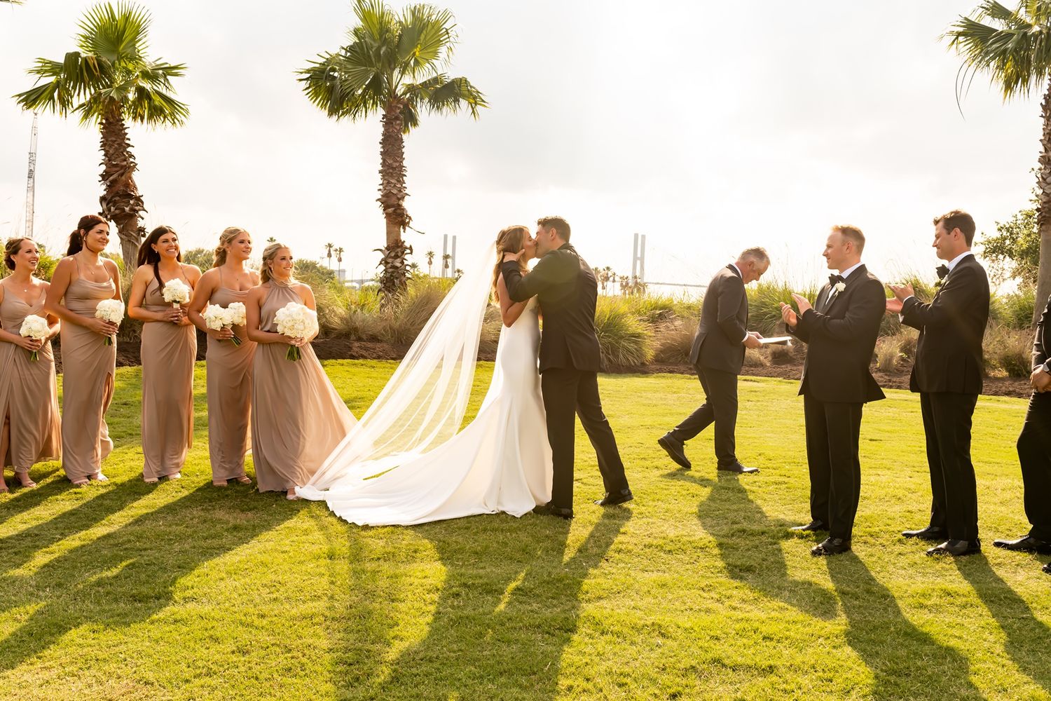 Wedding party standing in a line on a grassy field with palm trees silhouetted against golden sunlight.