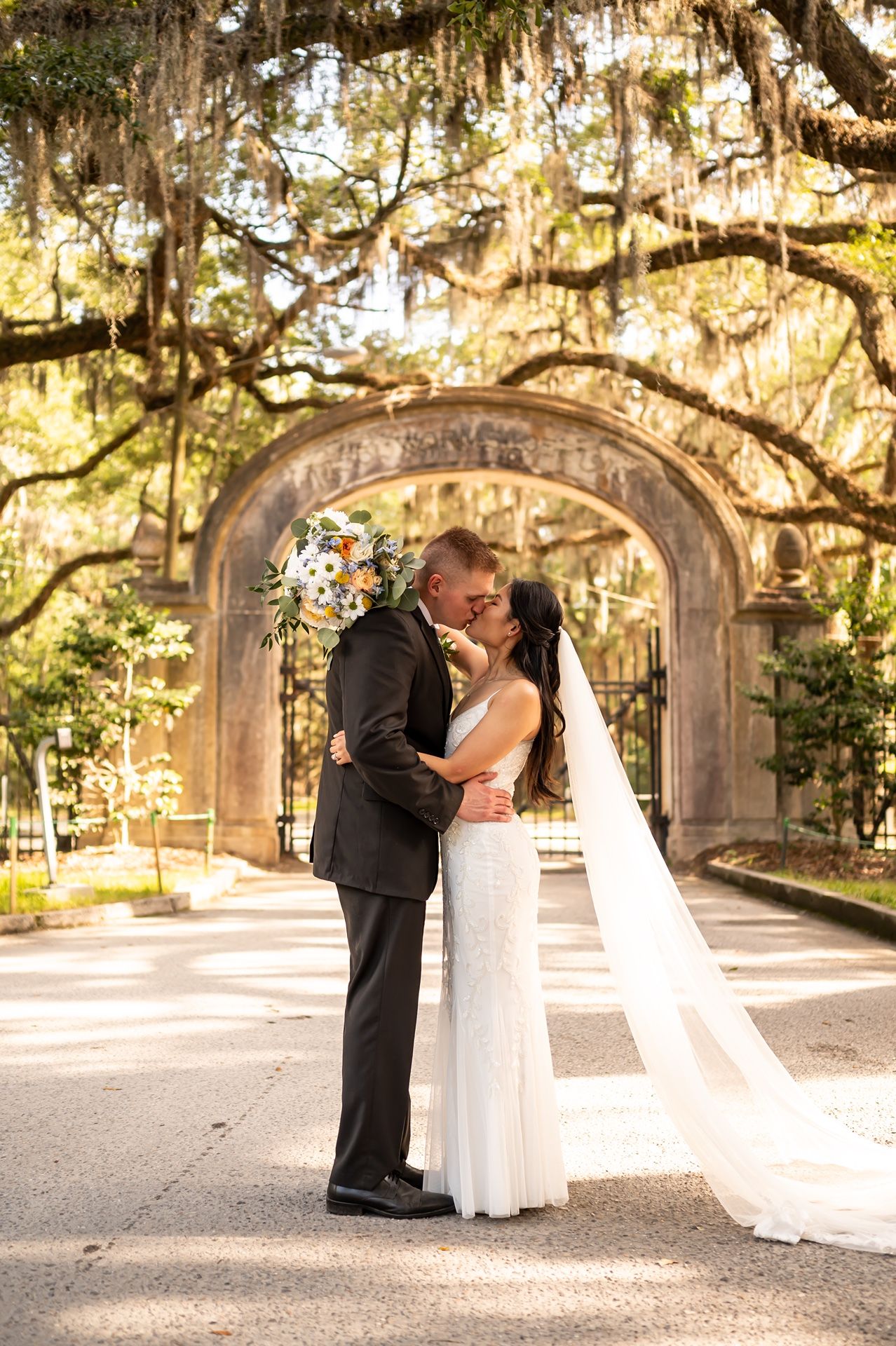 A romantic newlywed couple shares a kiss under a Spanish moss-draped stone archway in a scenic southern garden.