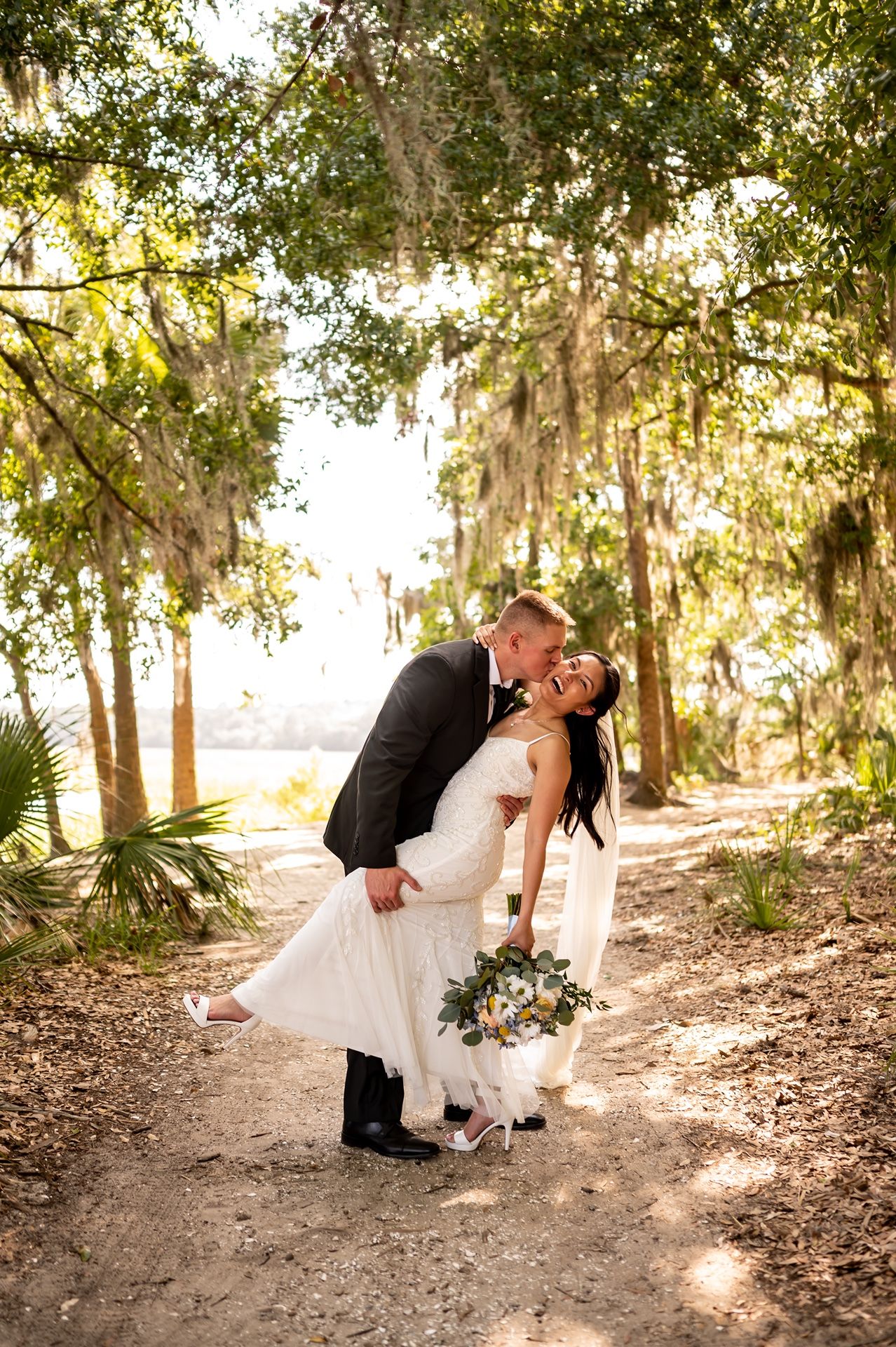 A romantic wedding couple shares an intimate dip pose in a sunlit wooded forest setting.