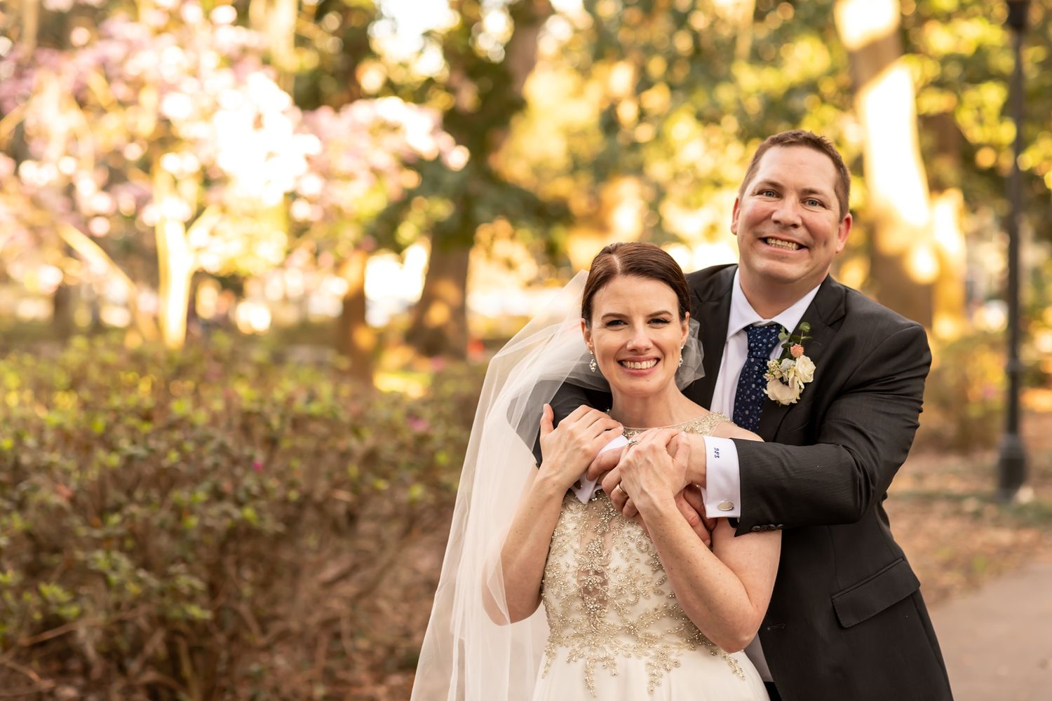 A couple wearing wedding attire embrace and smile while standing on an autumn path with golden trees in the background.