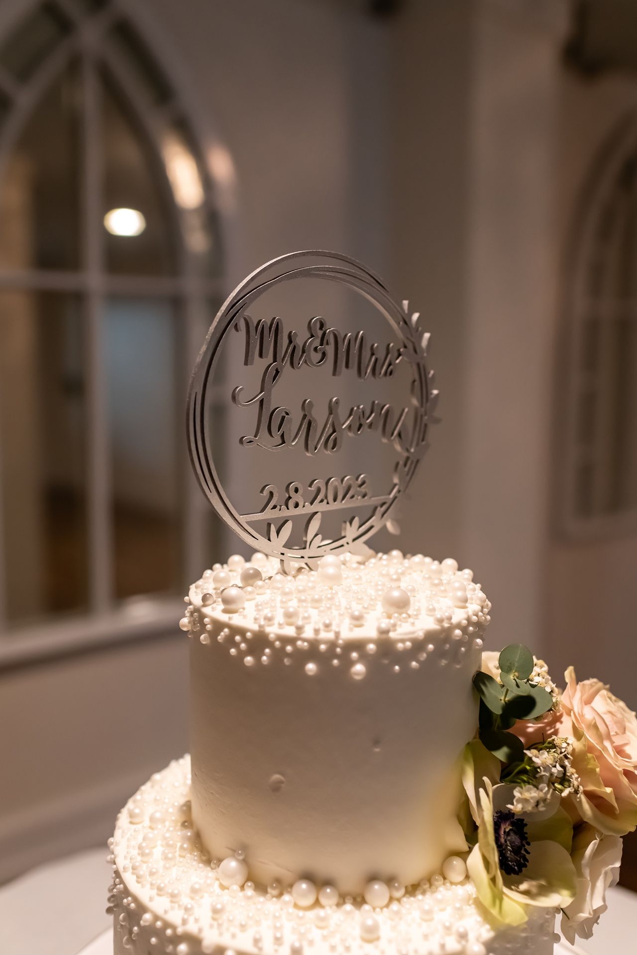A white wedding cake decorated with delicate flowers and a circular metallic cake topper illuminated by warm lighting.