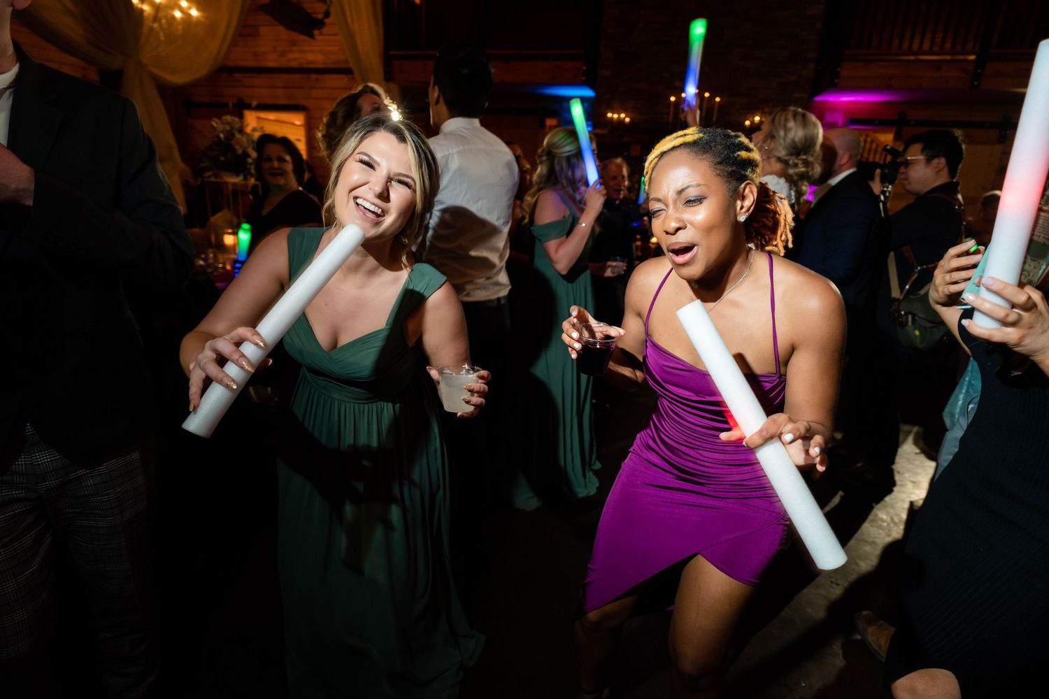 Partygoers in green and purple dresses dance energetically with glowing foam sticks at a nightclub celebration.