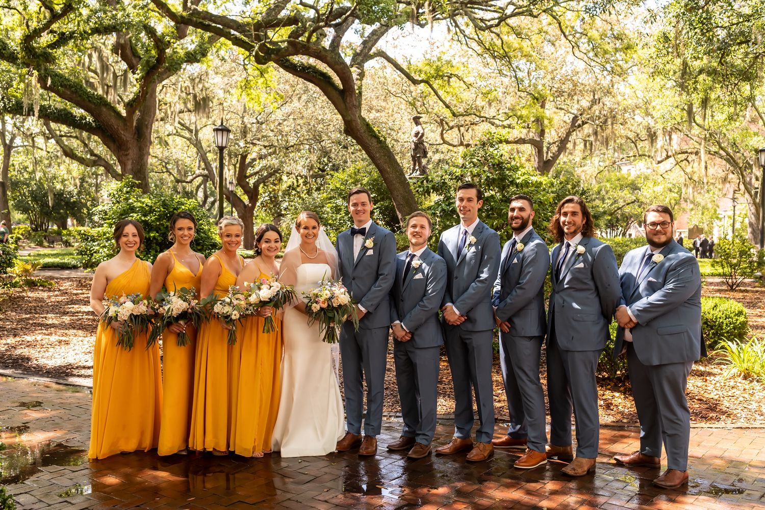 Wedding party in yellow and gray attire poses under majestic oak trees during outdoor celebration.