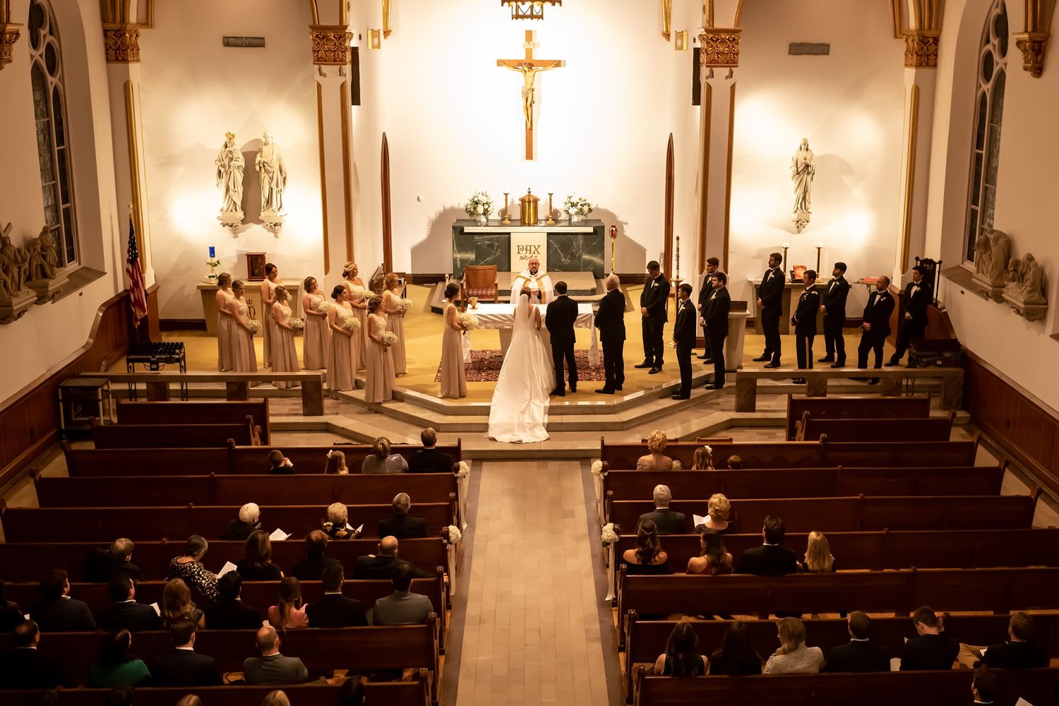 Church interior with wooden pews shows ceremony attendees gathering in rows beneath ornate religious decor and lighting.