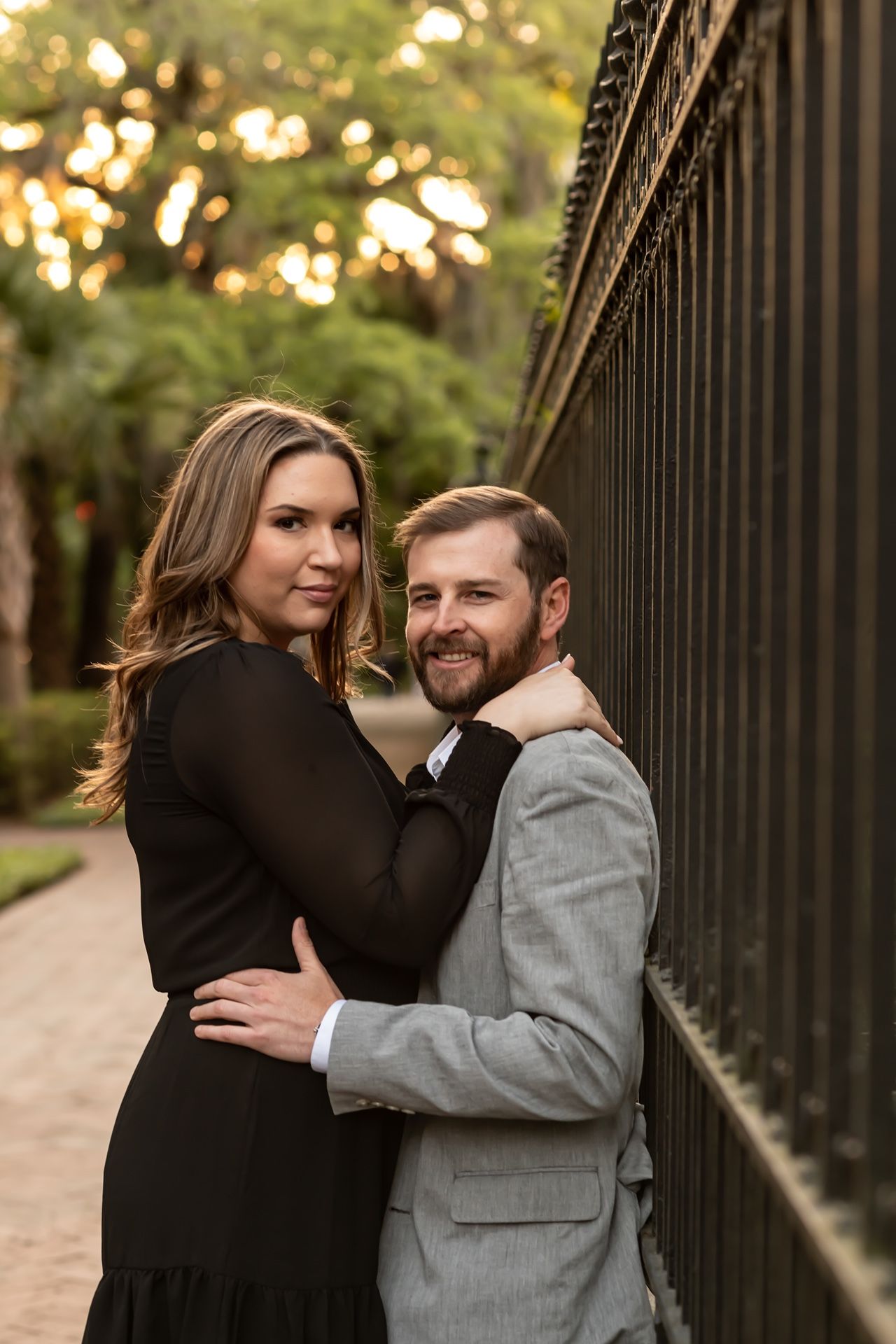 Romantic couple poses against iron fence at sunset with bokeh lighting in outdoor park setting.