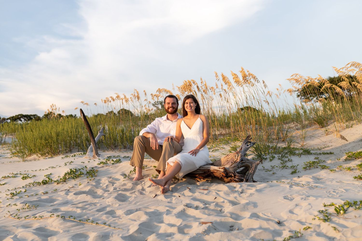People gather on a sandy beach at sunset, surrounded by sea grass and natural coastal landscape.