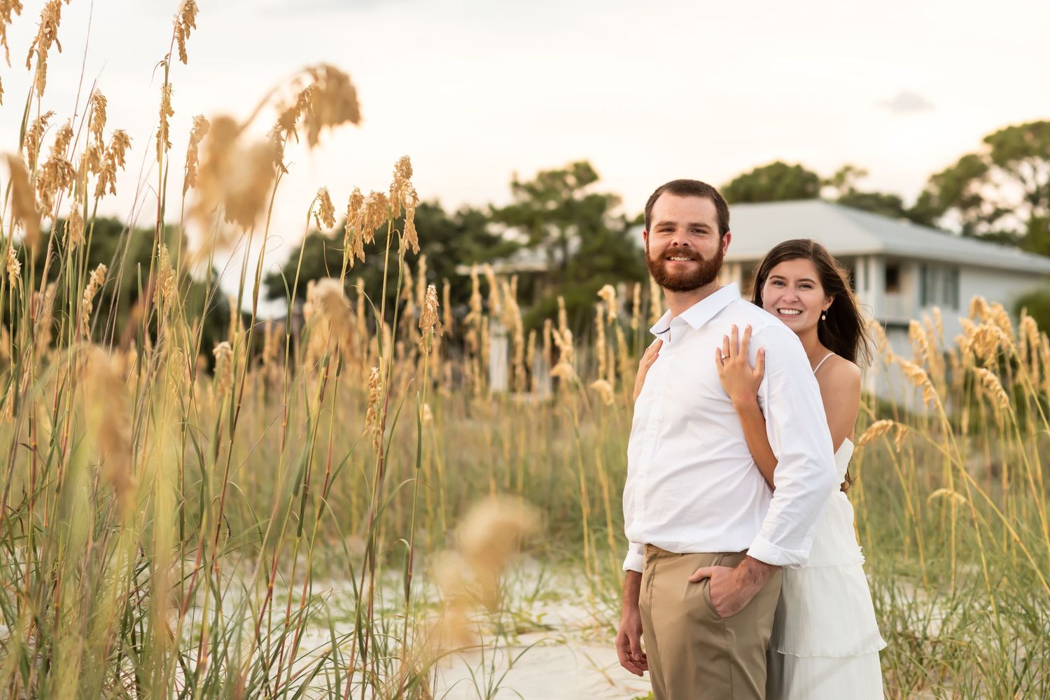 A couple stands lovingly together among tall beach grass during golden hour near a coastal house.