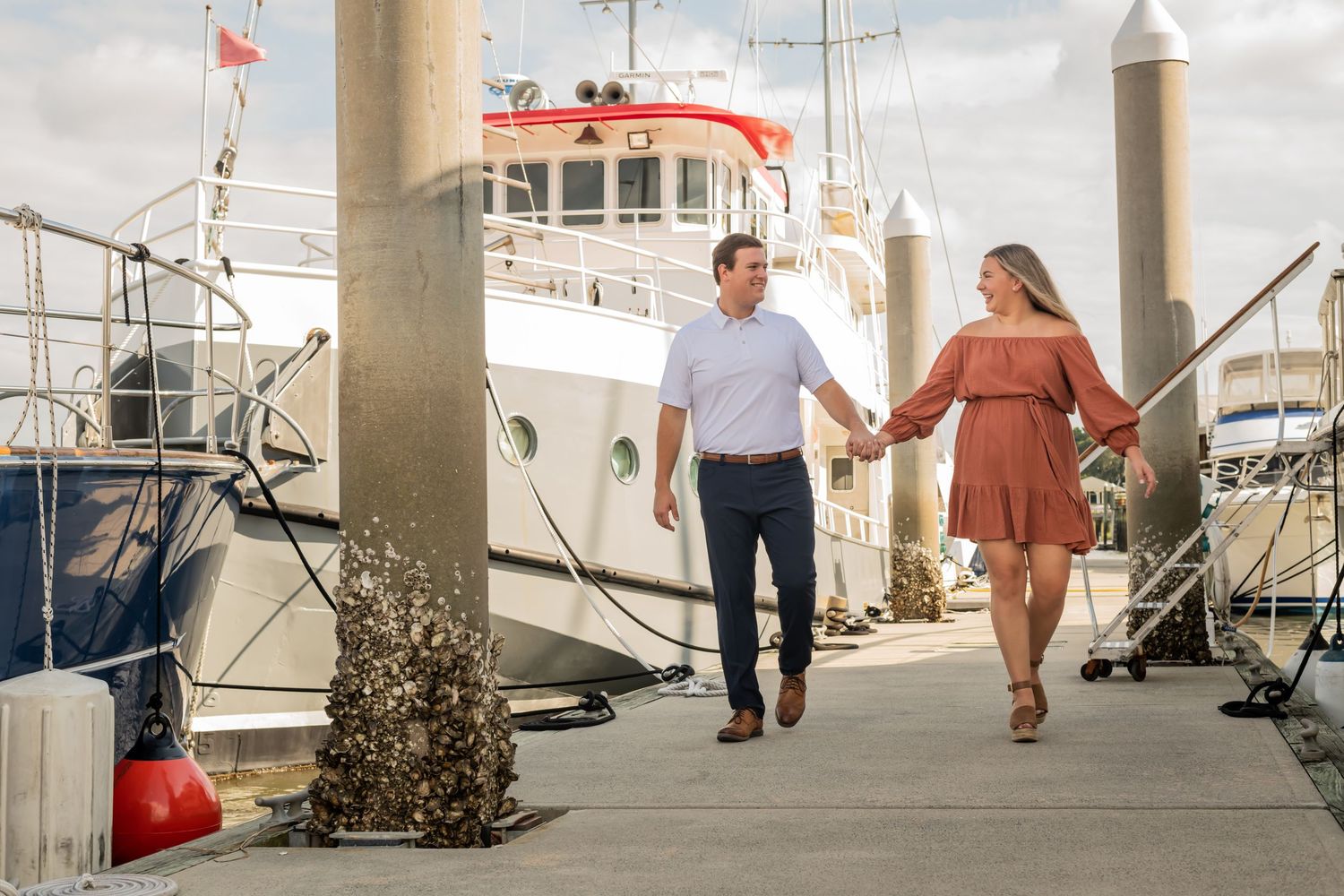 A couple holding hands walks along a marina dock with boats and nautical equipment visible in the background.