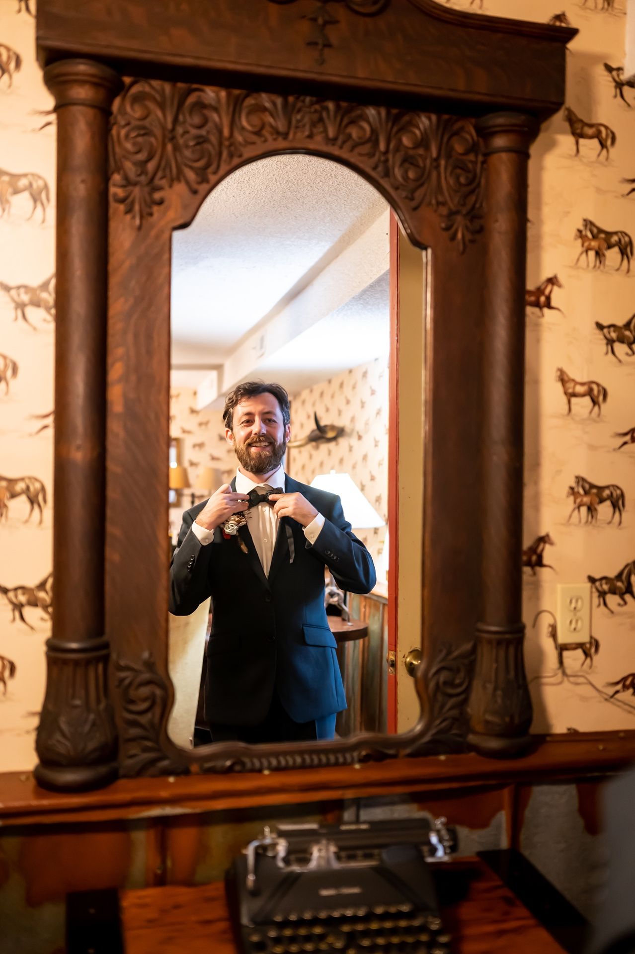 Reflection in an ornate wooden mirror showing someone adjusting their bowtie, with vintage wallpaper and typewriter visible.
