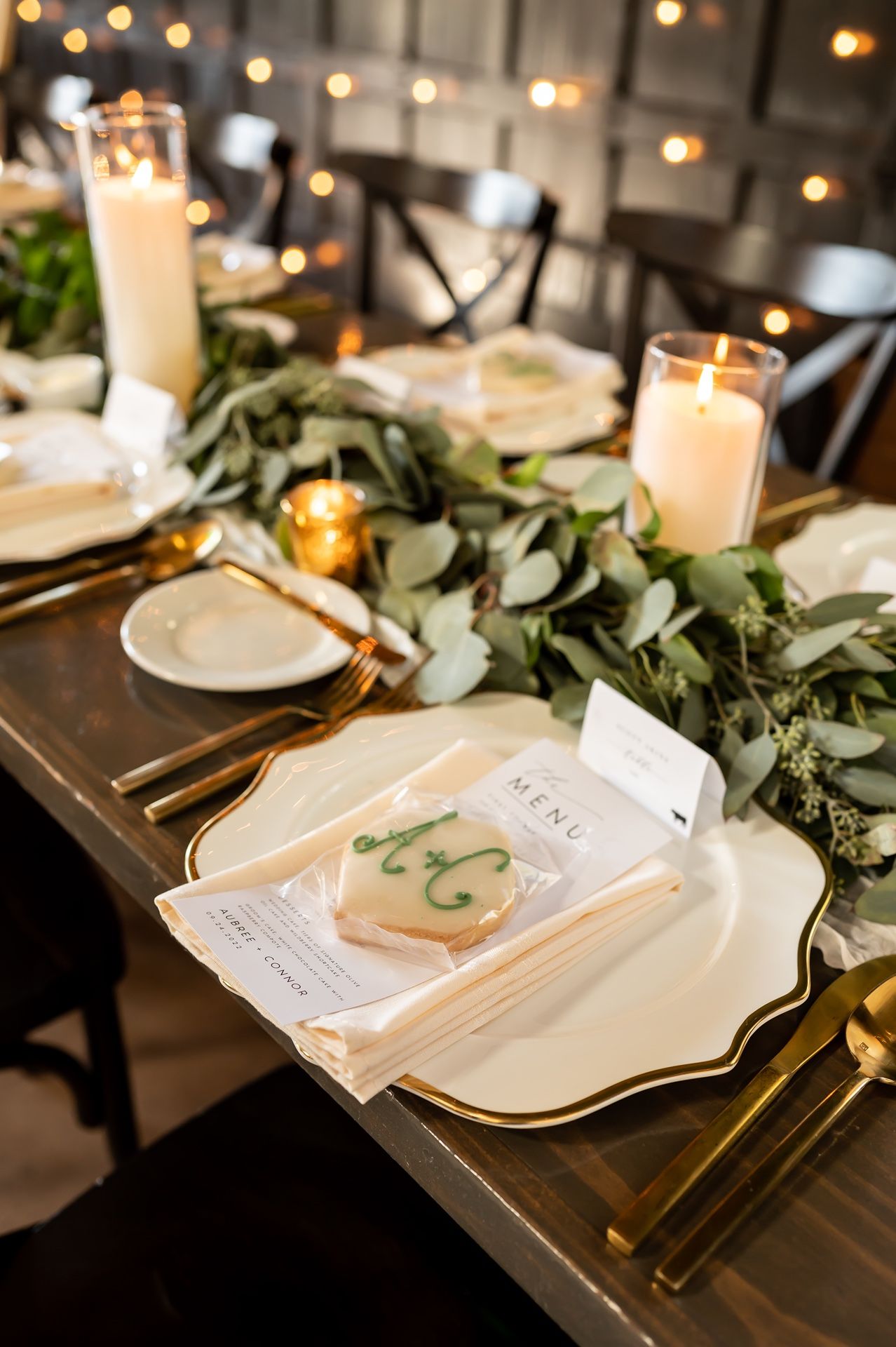 Elegant wedding table place setting with gold flatware, white plates, and eucalyptus greenery against dark wood.