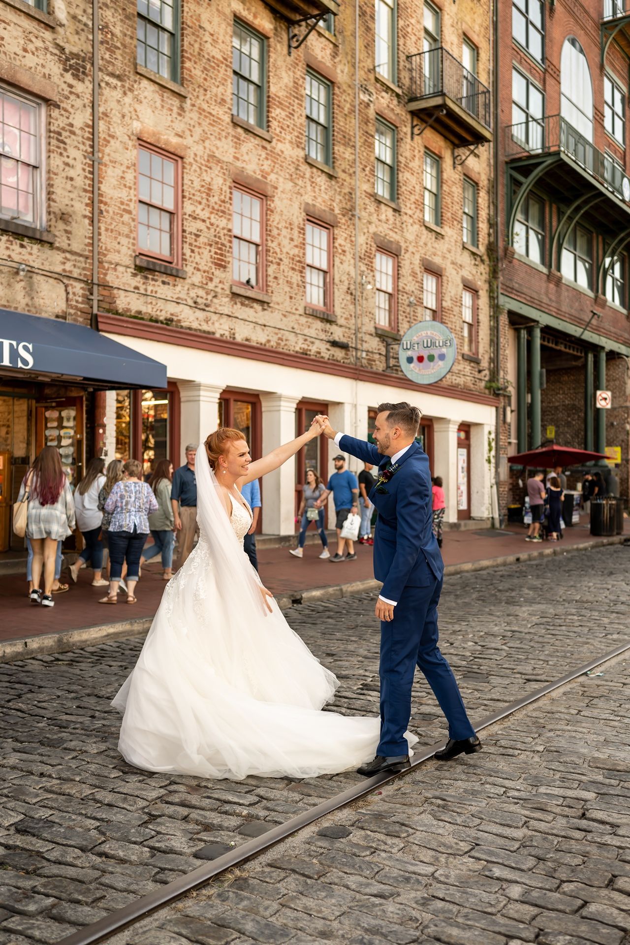 A wedding couple dances on a historic cobblestone street with brick buildings and shops in the background.