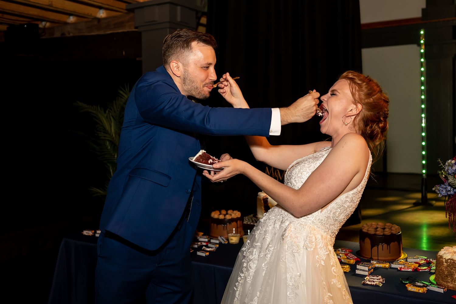 Two people in wedding attire playfully feed each other cake during a reception celebration in an indoor venue.