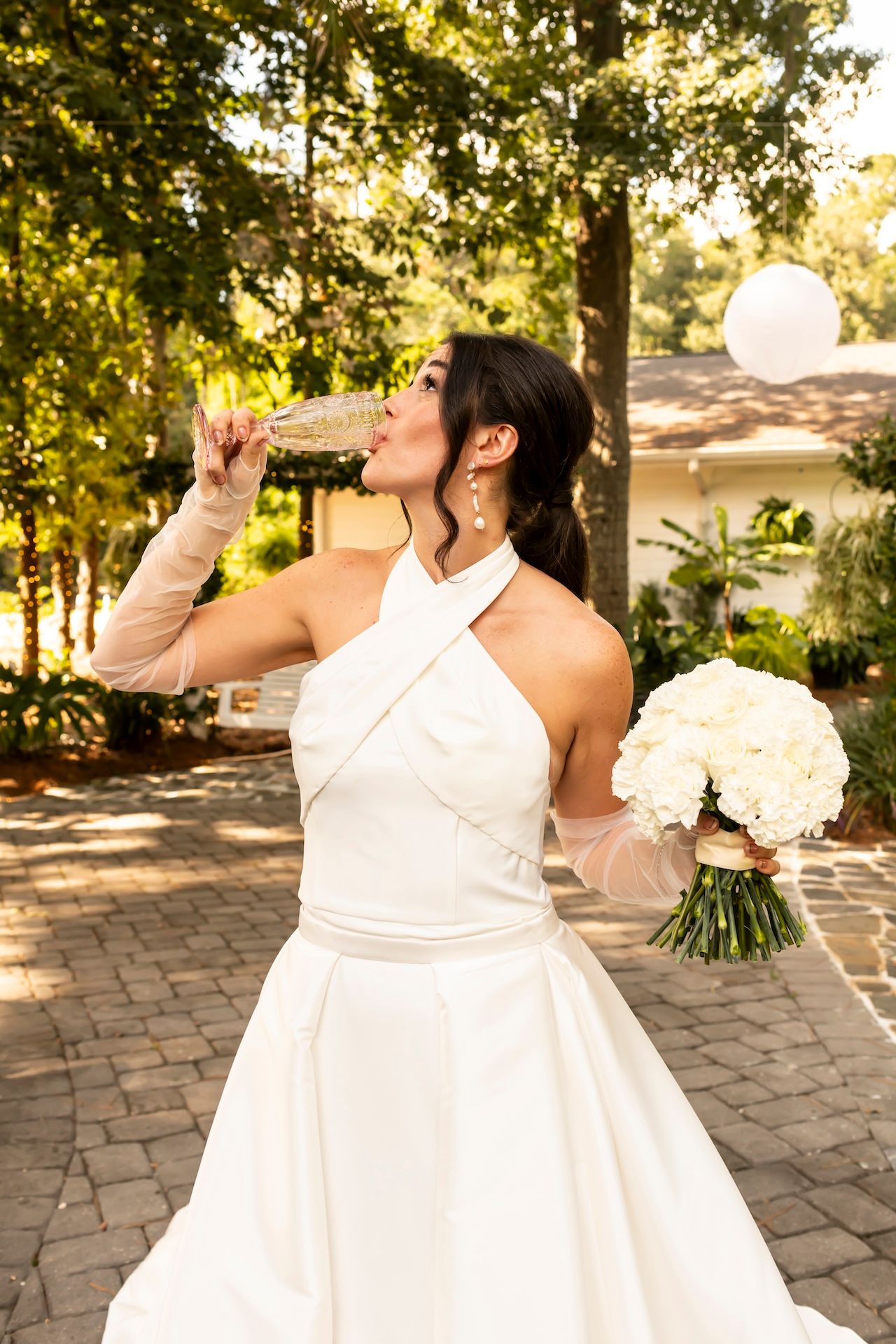 A bride in a halter neck wedding gown drinks from a bottle while holding a white bouquet on a brick patio.