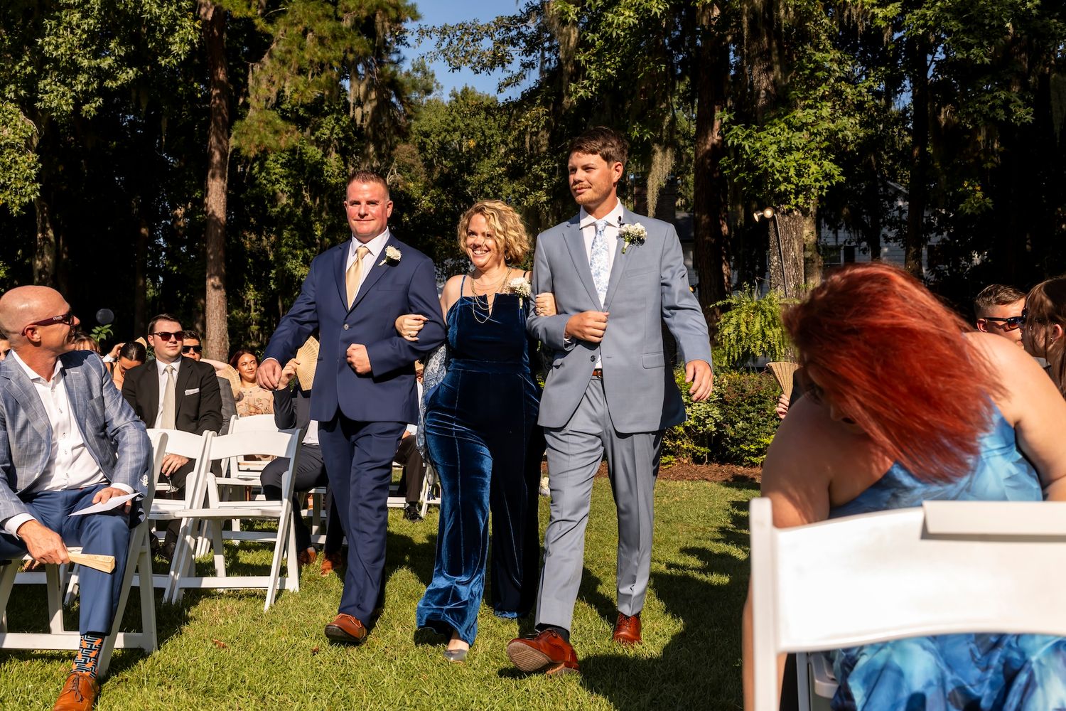 Wedding attendees walk down the outdoor aisle during a garden ceremony surrounded by trees and white chairs.
