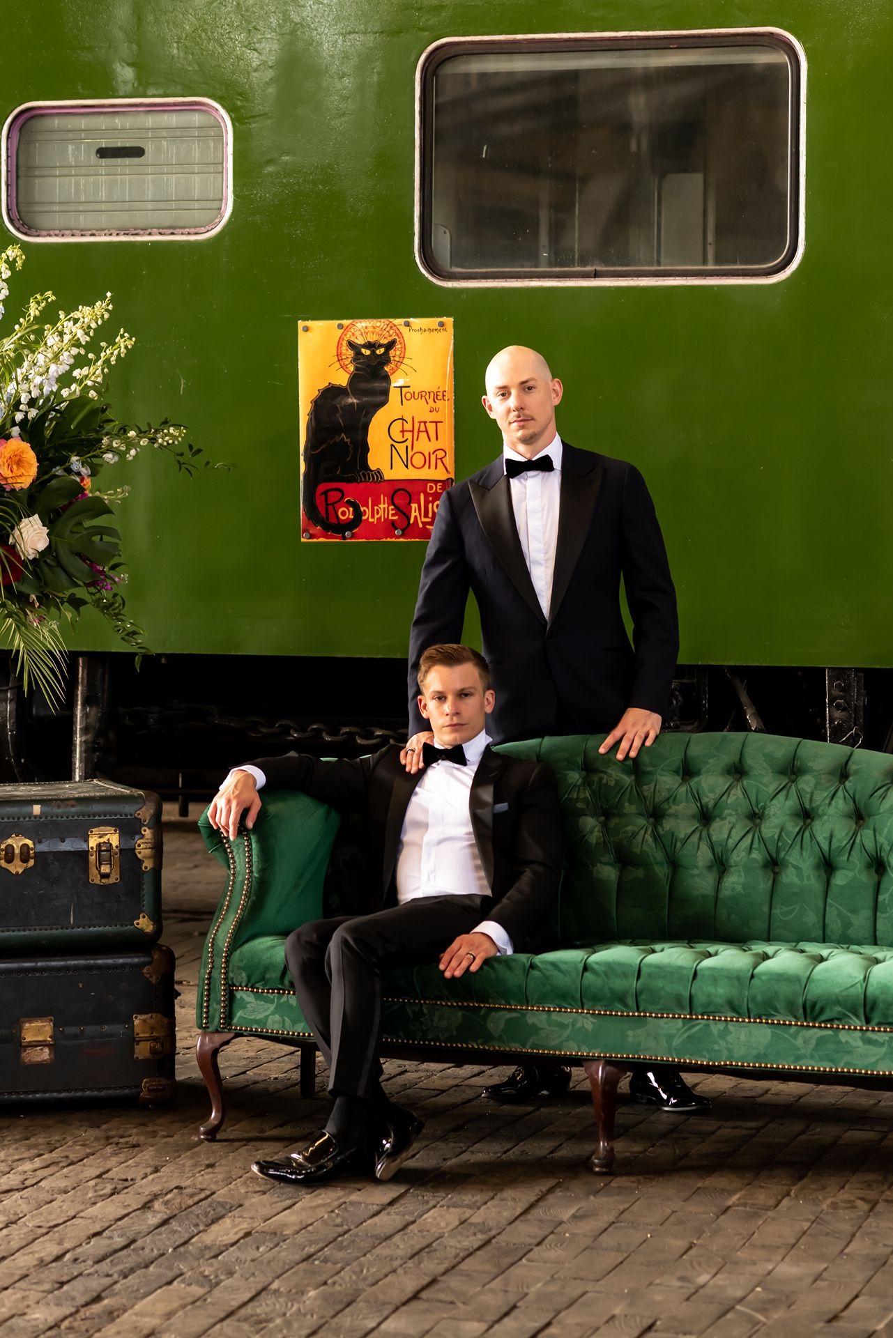Two people in formal black tuxedos pose by a vintage green sofa with a black cat poster on green wall behind them.