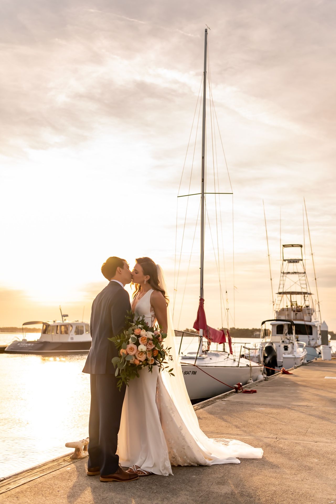 A loving couple shares a romantic moment on a marina dock at sunset with boats in the background.