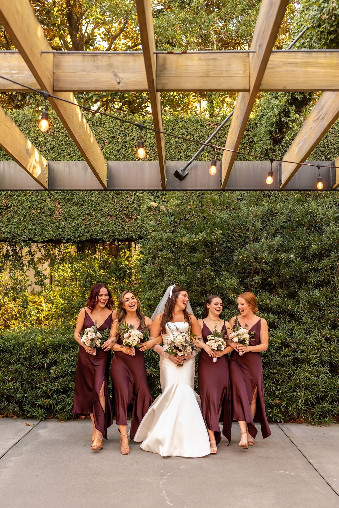 Wedding party stands under wooden pergola with greenery backdrop wearing burgundy dresses at outdoor venue.