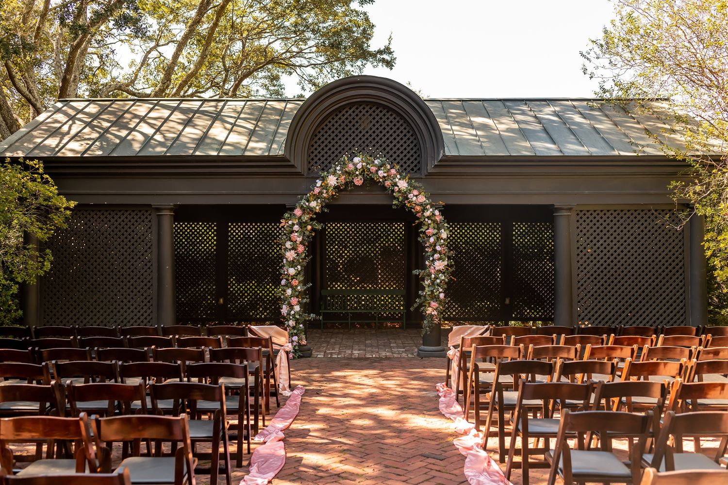 Outdoor wedding venue setup with wooden chairs and floral arch for ceremony in natural lighting.