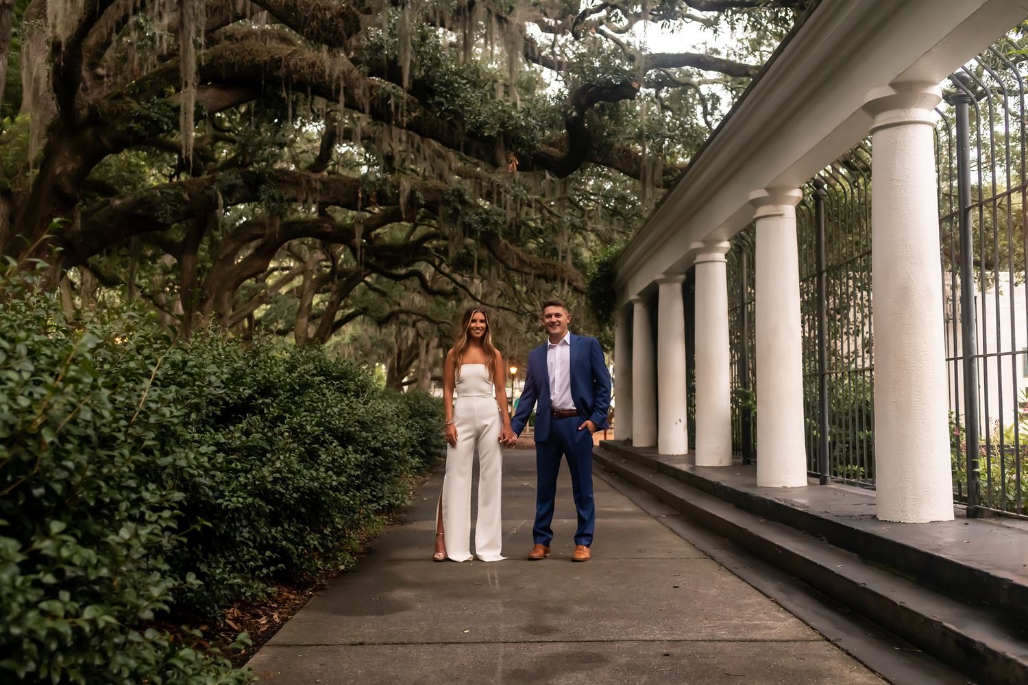 A couple walks together along a covered porch with white columns surrounded by lush greenery on a cloudy day.