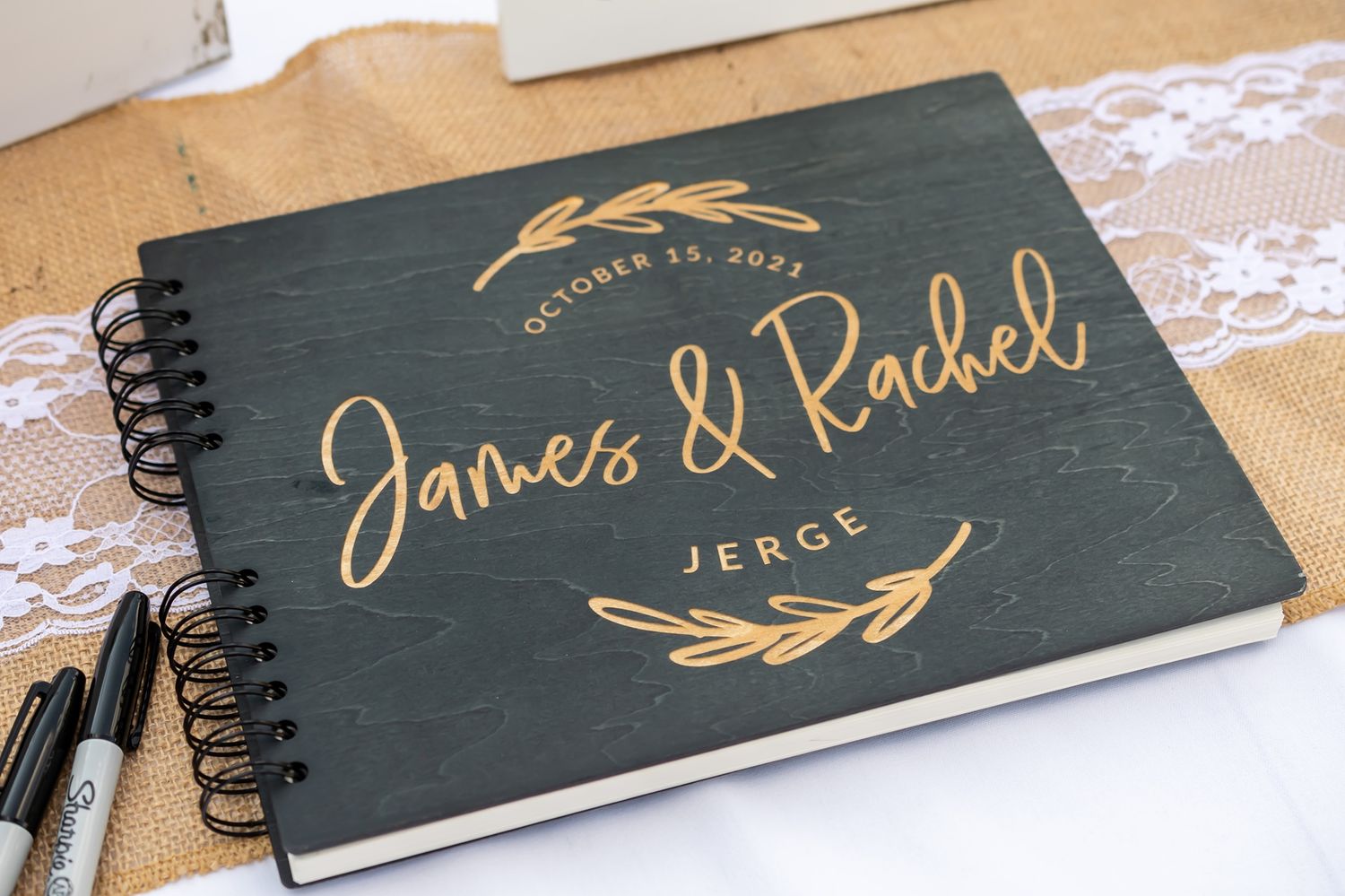 Black hardcover guestbook with elegant gold calligraphy text and decorative leaf design on wooden table with lace detail.