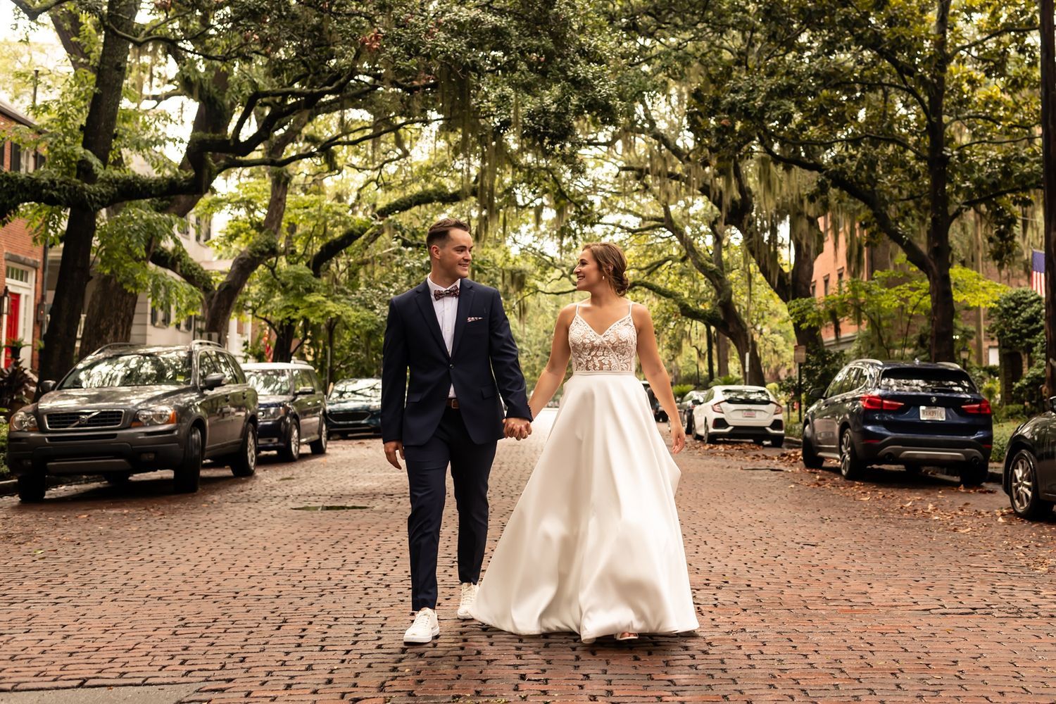 A couple walking down a tree-lined cobblestone street in their wedding attire on a beautiful autumn day.