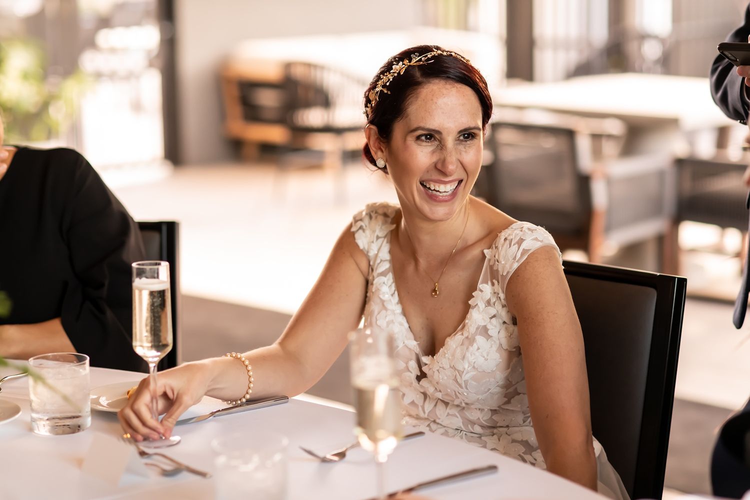 A person wearing a white lace wedding dress smiles while seated at an elegant restaurant table.
