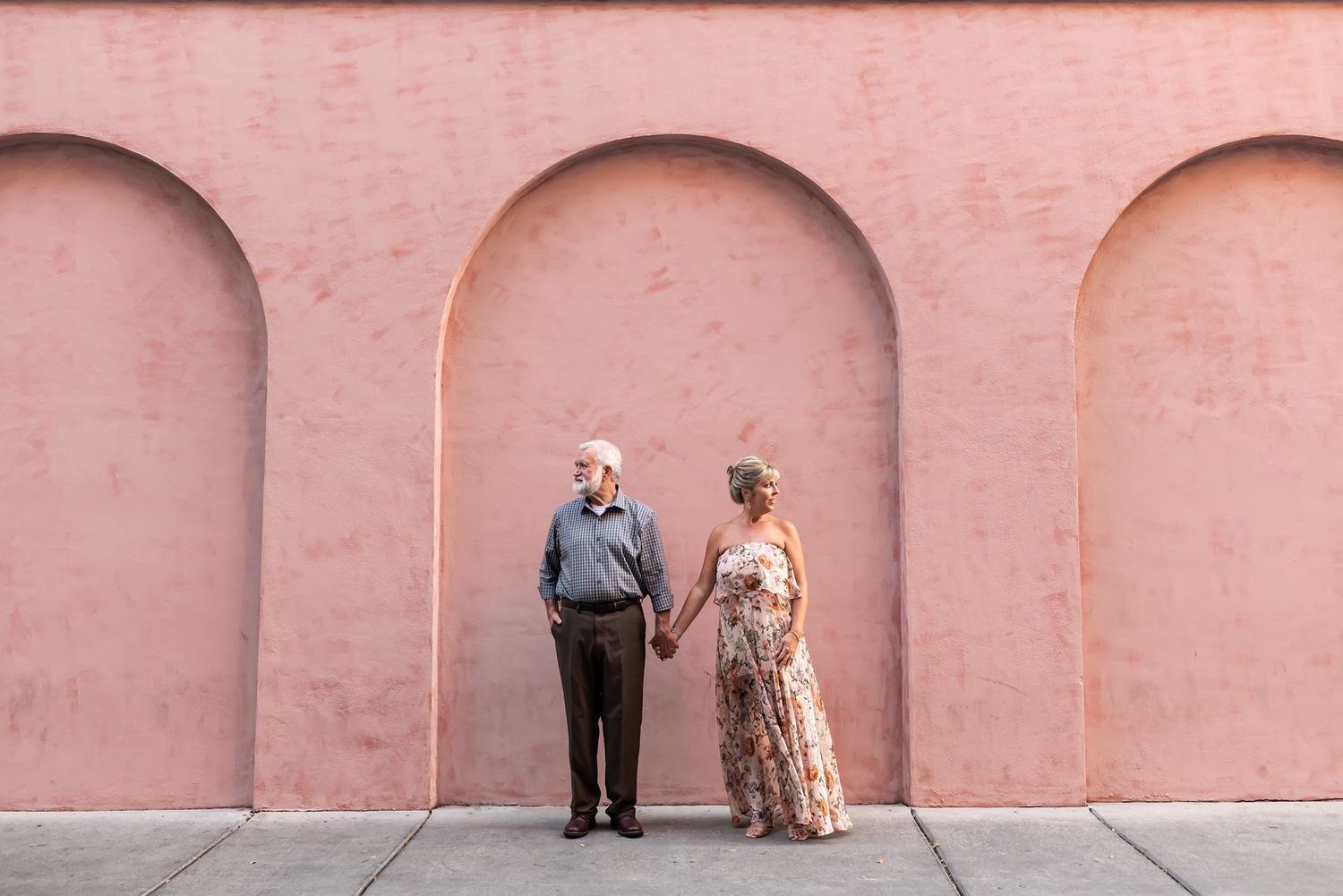 A stylishly dressed couple poses against a pink wall with arched recesses in Charleston.