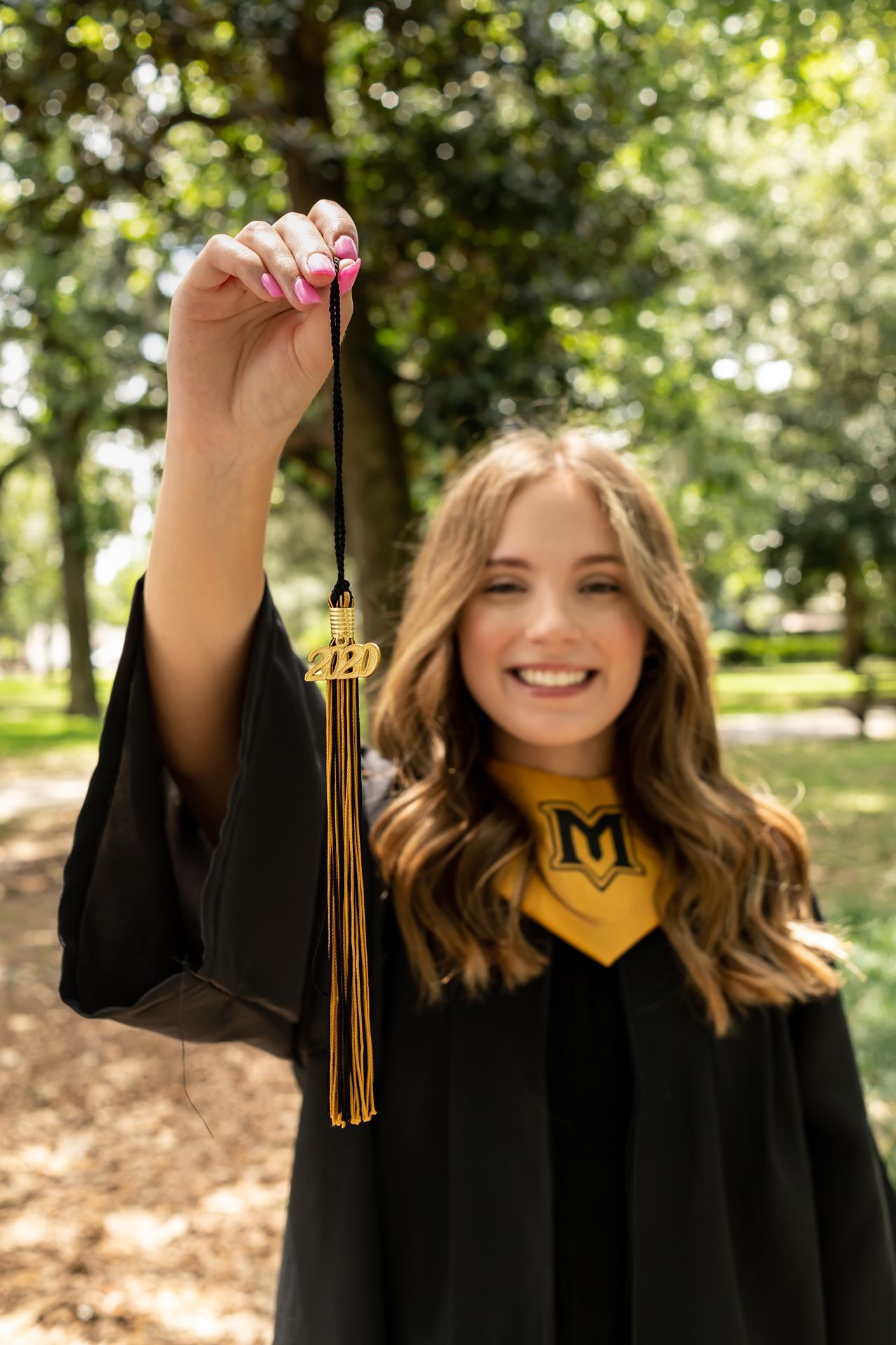 Graduate in black gown displays tassel with bright smiles in park setting.