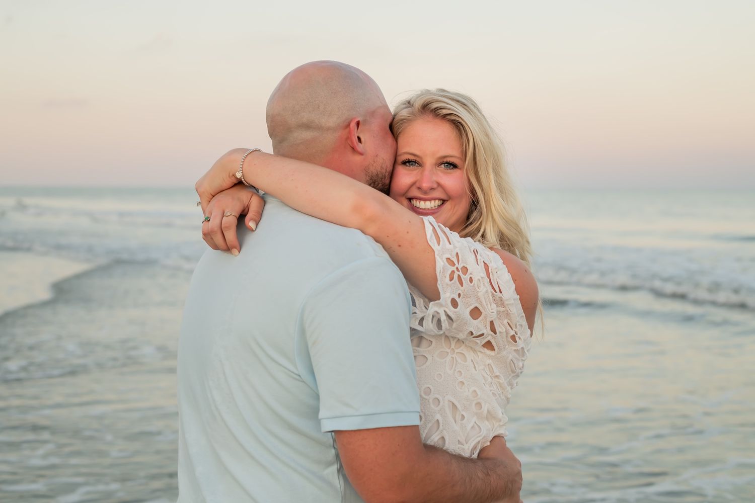 A couple embraces on the beach at sunset with ocean waves rolling in behind them as they share a joyful moment together.