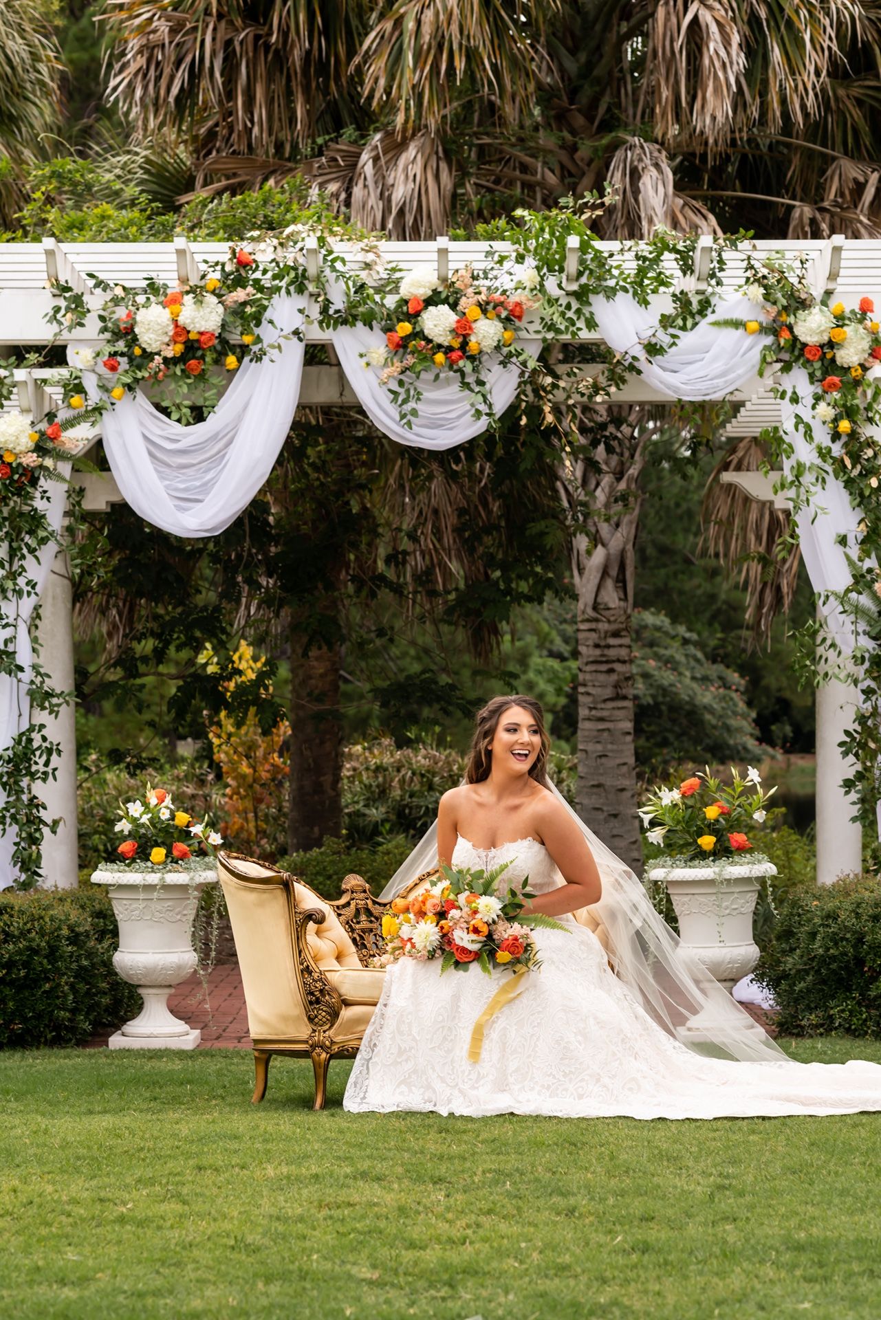 Wedding portrait under decorated white pergola with orange and white floral arrangements and draped fabric.