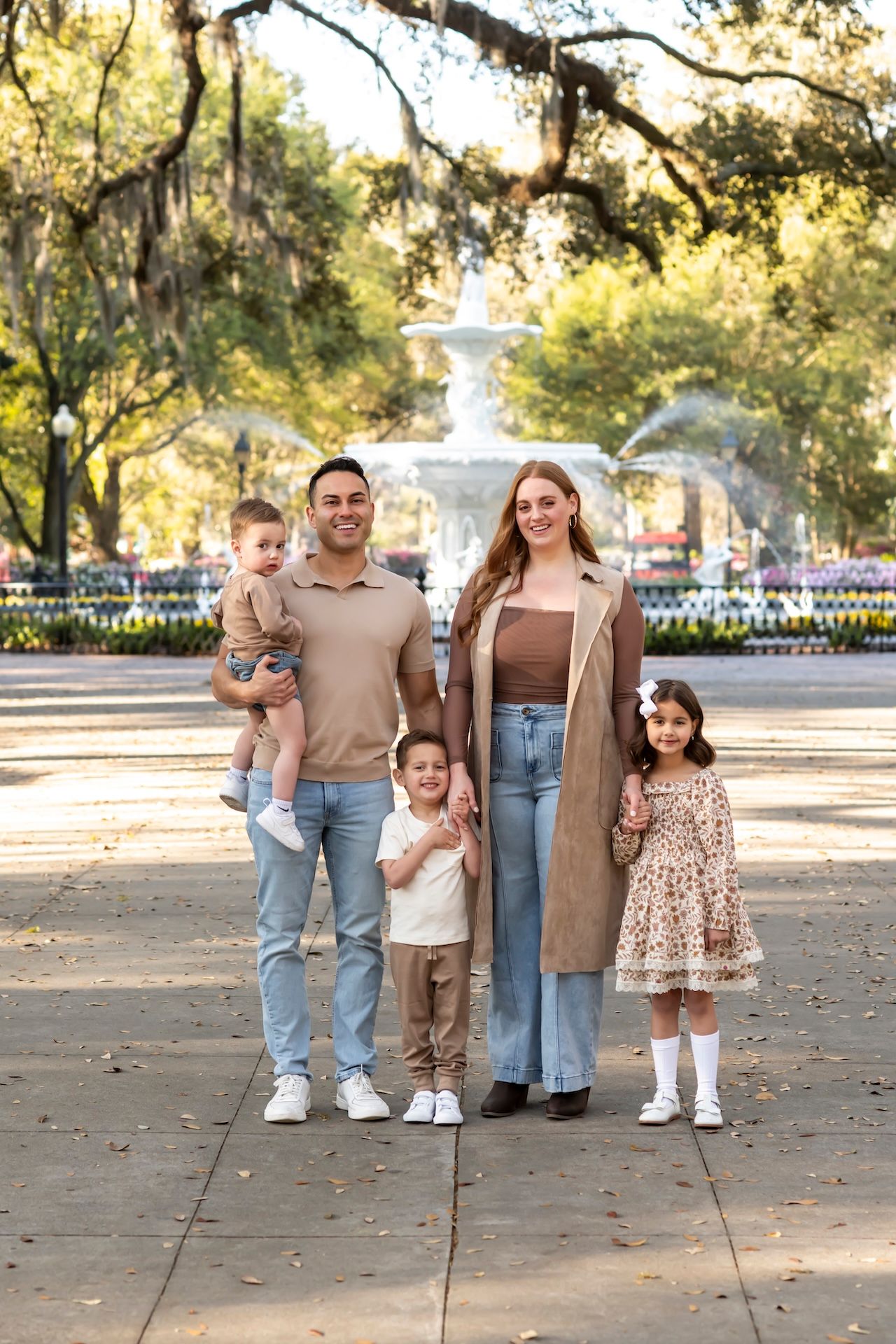 Family with three children standing on a tree-lined path in a park with a white gazebo in the background.