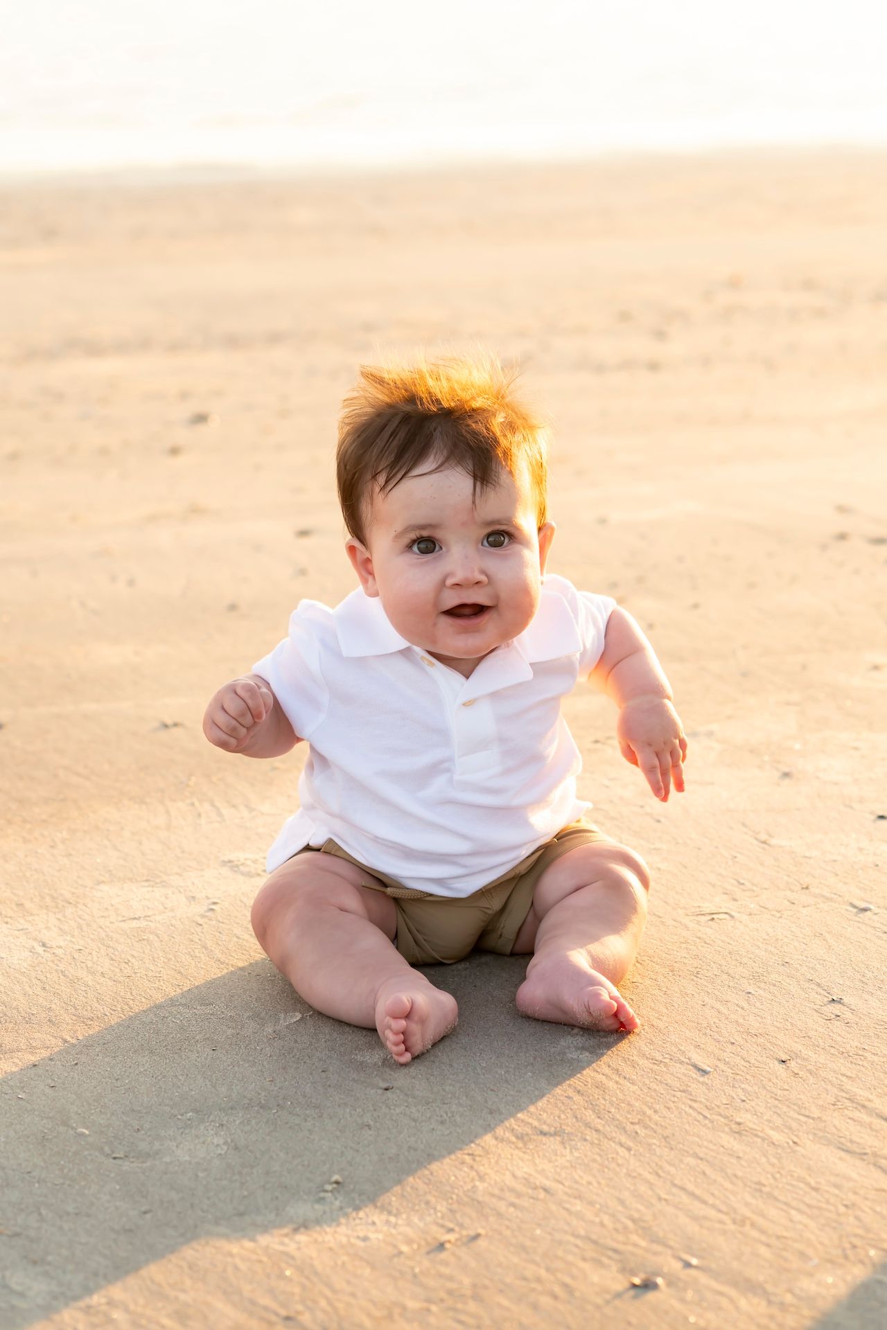 Cheerful baby wearing white polo and khaki shorts sits on sandy beach at golden hour.
