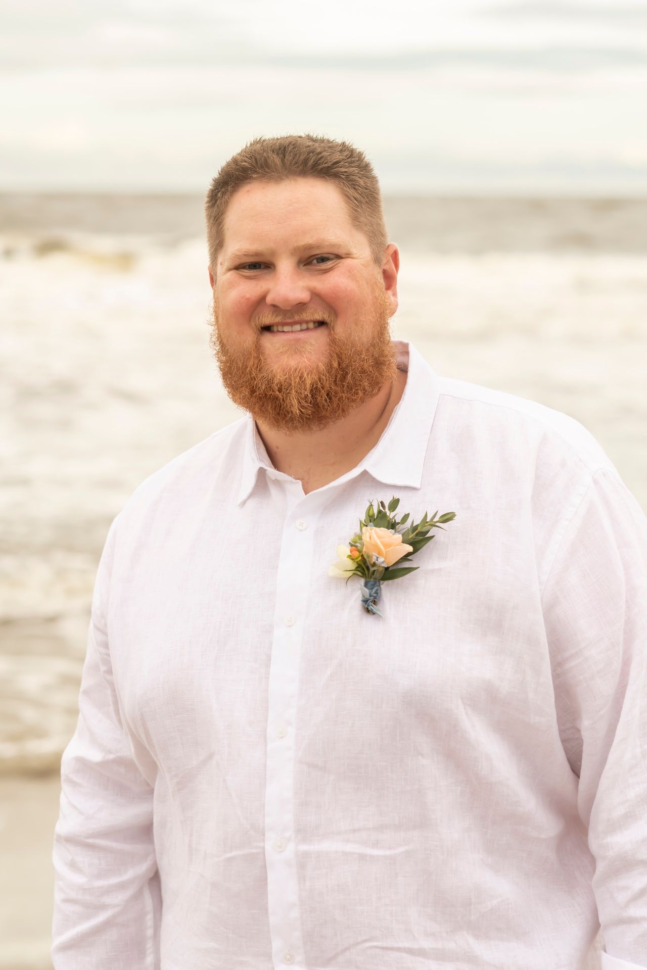 A groom in a white dress shirt with a boutonniere smiles on the beach during a wedding photoshoot.