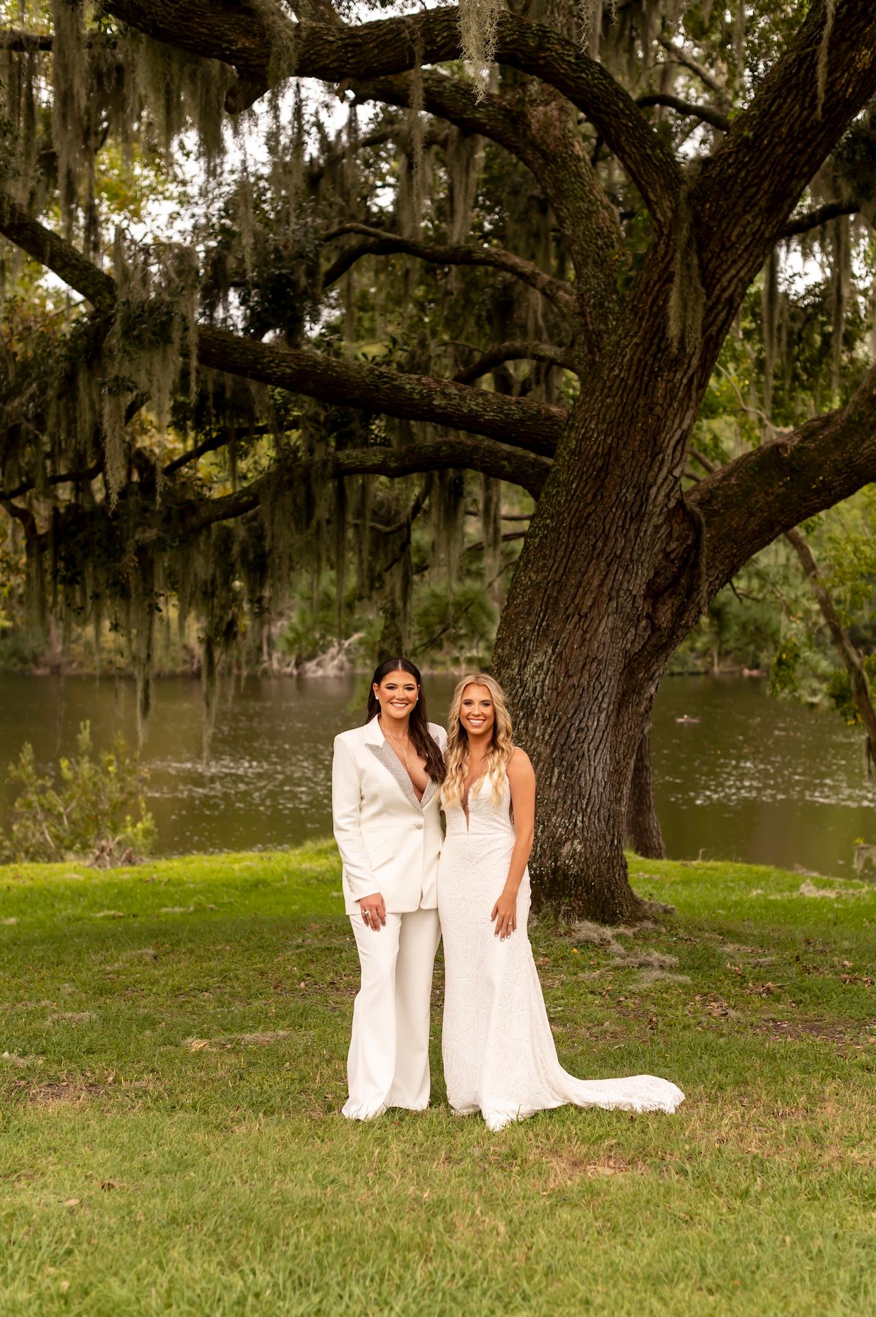 Two people wearing white dresses embrace while standing on grassy grounds under mossy oak trees near a lake.